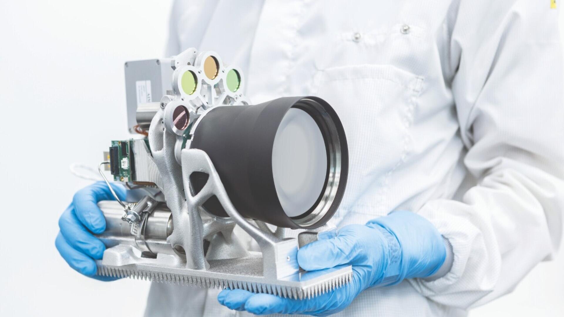 A person in a white lab coat and blue gloves holds a large, advanced scientific camera or optical instrument with multiple lenses and electronic components attached.