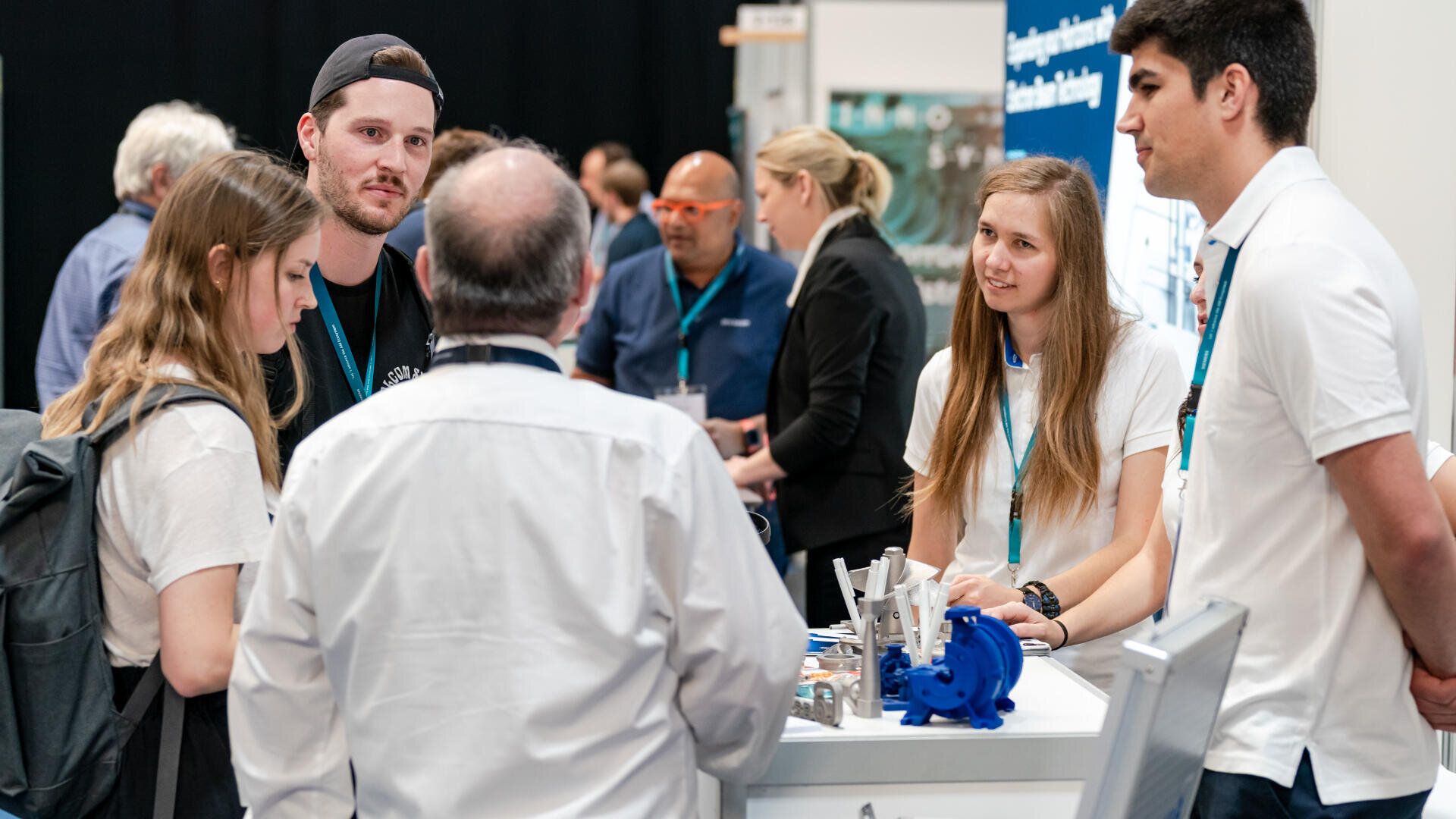 Eine Gruppe junger Erwachsener unterhält sich während einer gut besuchten Veranstaltung oder Messe an einem Stand mit einem älteren Mann. Im Hintergrund sieht man Menschen, die sich unterhalten und Kontakte knüpfen.