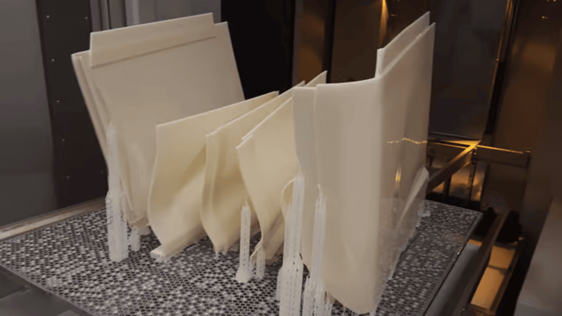 Mehrere große, cremefarbene 3D-gedruckte Komponenten stehen aufrecht auf einer strukturierten Plattform in einem 3D-Drucker, wobei an einigen Teilen sichtbare Stützstrukturen angebracht sind.