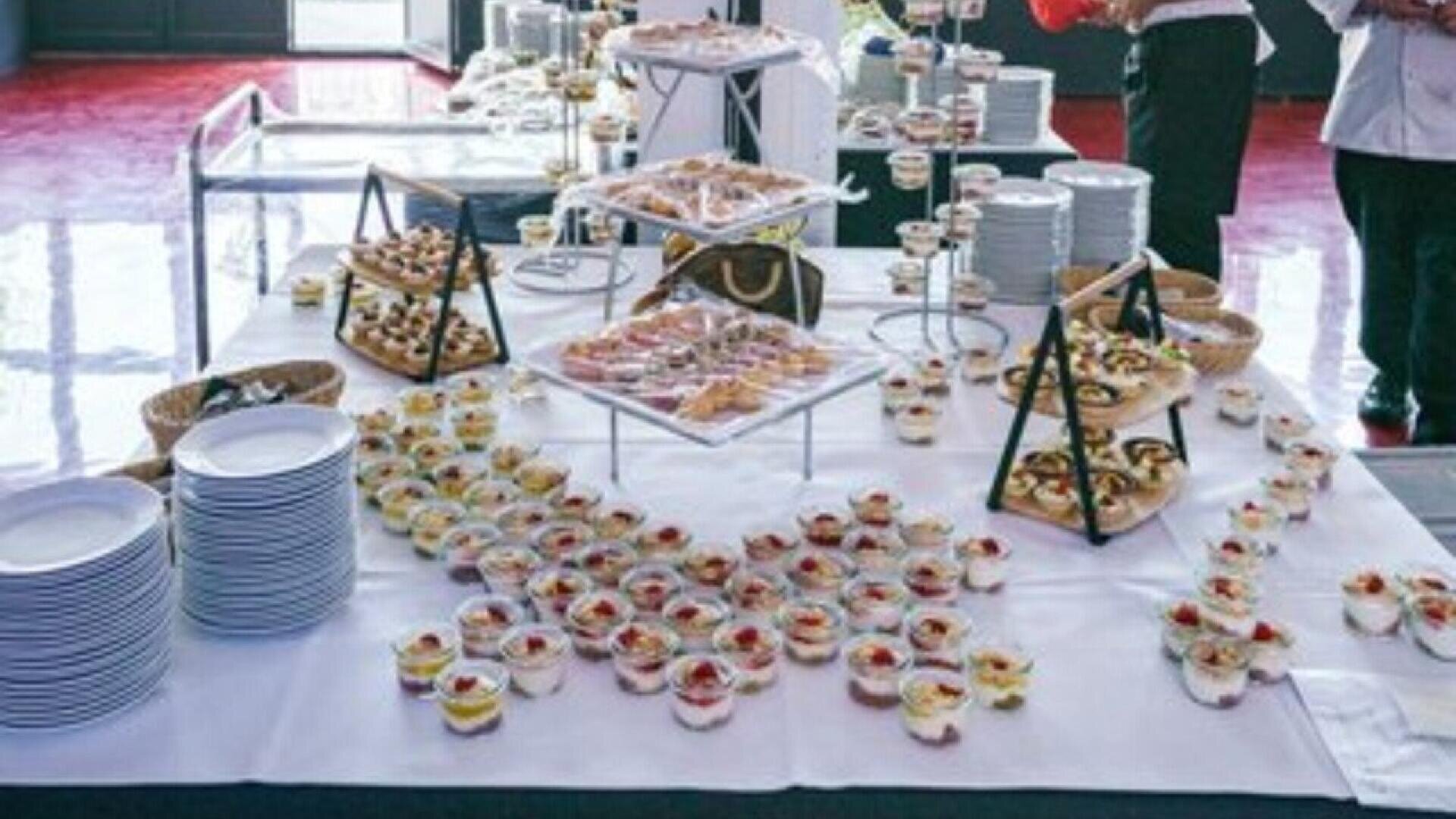 A buffet table covered with a white tablecloth displays an assortment of small desserts and appetizers on trays and tiered stands, with stacks of white plates and people standing nearby.