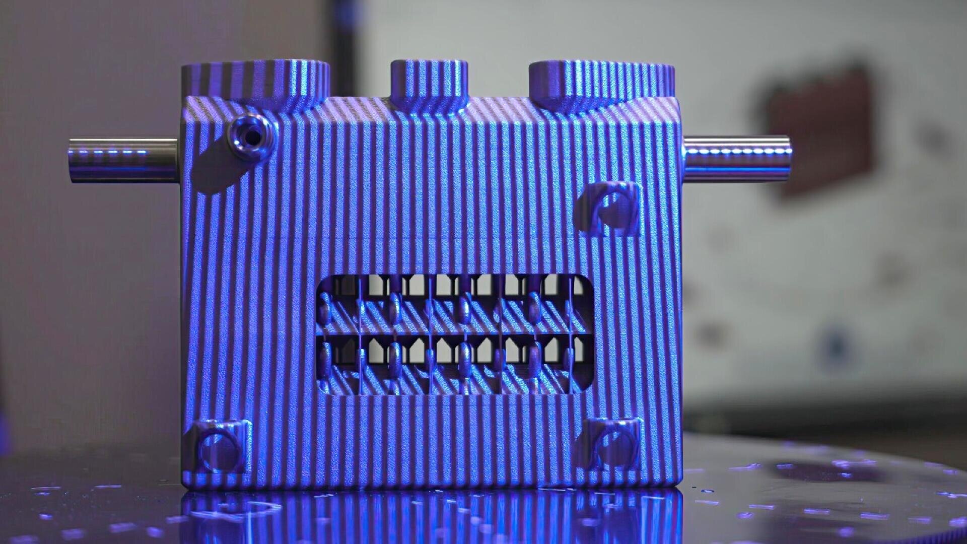 A metal mechanical device with vertical grooves, bolts at each corner, a horizontal rod passing through the top, and an open window showing internal components, all illuminated by blue light.