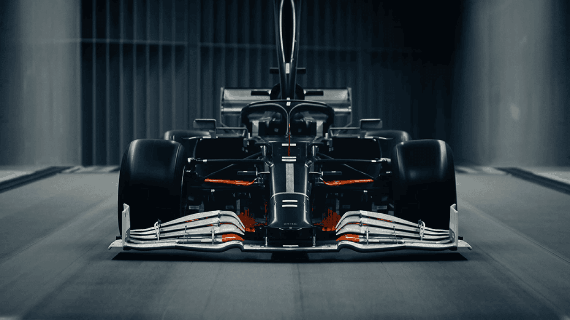 Front view of a sleek, black Formula 1 race car in a wind tunnel, with aerodynamic components and glowing orange lights highlighting details.
