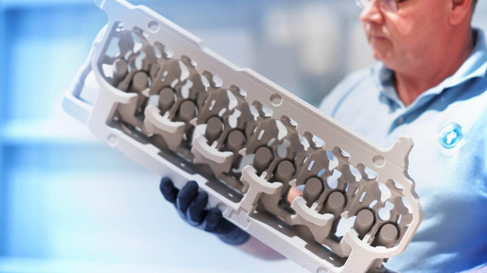 A person wearing glasses and gloves holds a large, intricate, metallic engine component, possibly a cylinder head, in a bright, industrial setting.