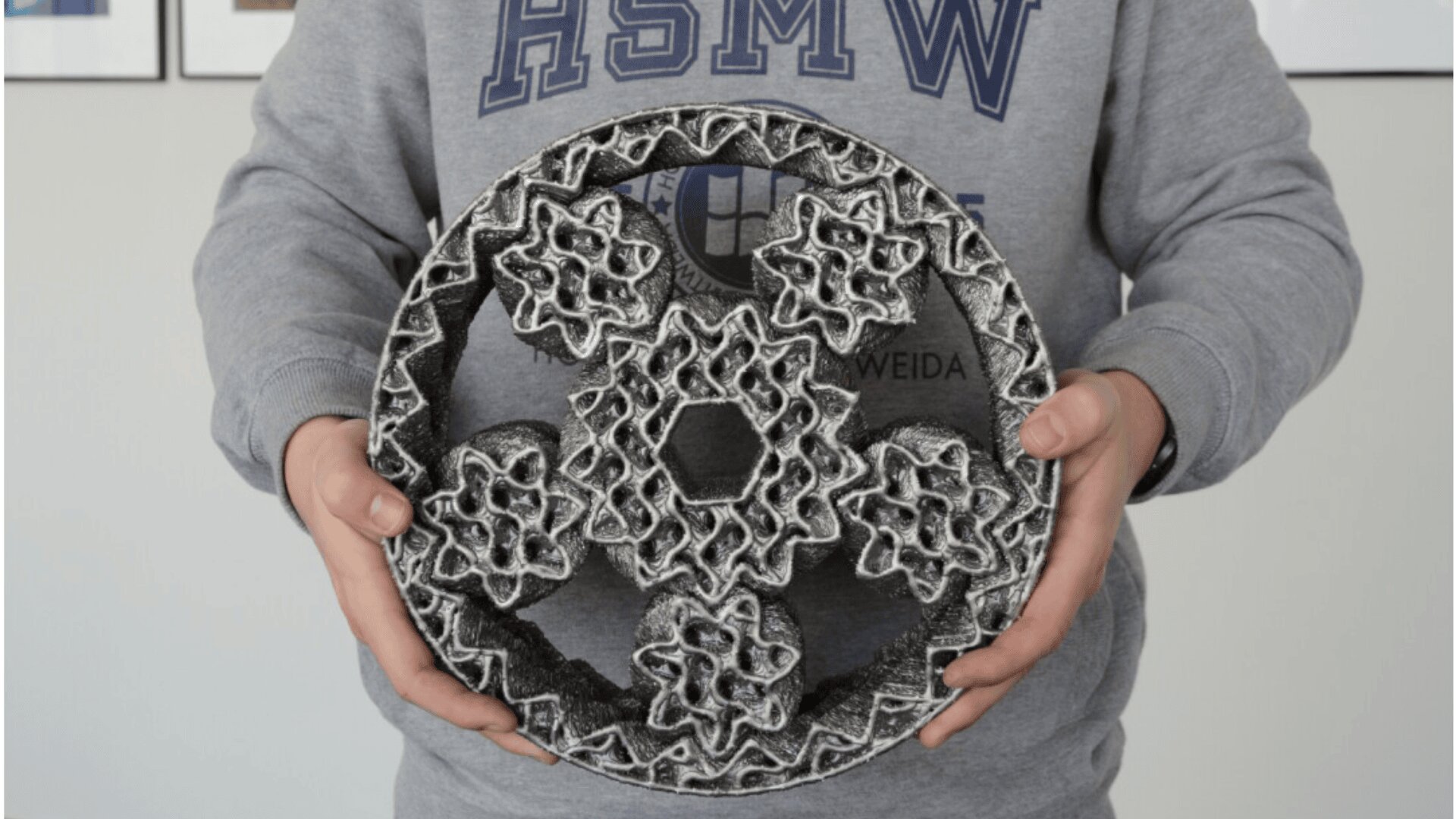 A person in a gray HSMW sweatshirt holds a large, intricate, gear-like 3D-printed object with interlocking geometric shapes in black and white.