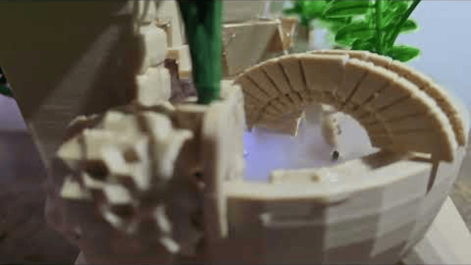 A close-up of a miniature water mill model with a small water wheel and an arched stone structure, surrounded by green plastic foliage.