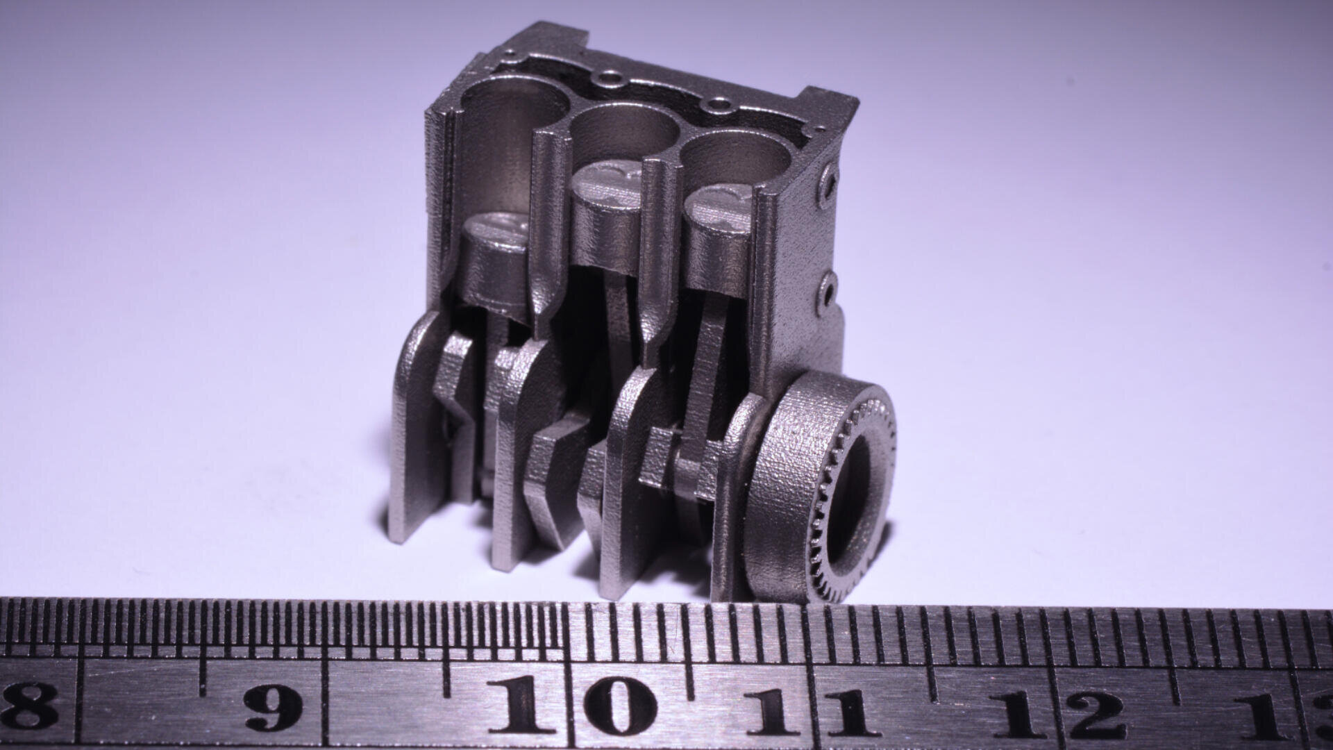Ein kleines, detailliertes mechanisches Metallteil, das einem Motor ähnelt, neben einem Zentimetermaß platziert, das auf einem weißen Hintergrund eine Höhe von etwa 3 cm anzeigt.