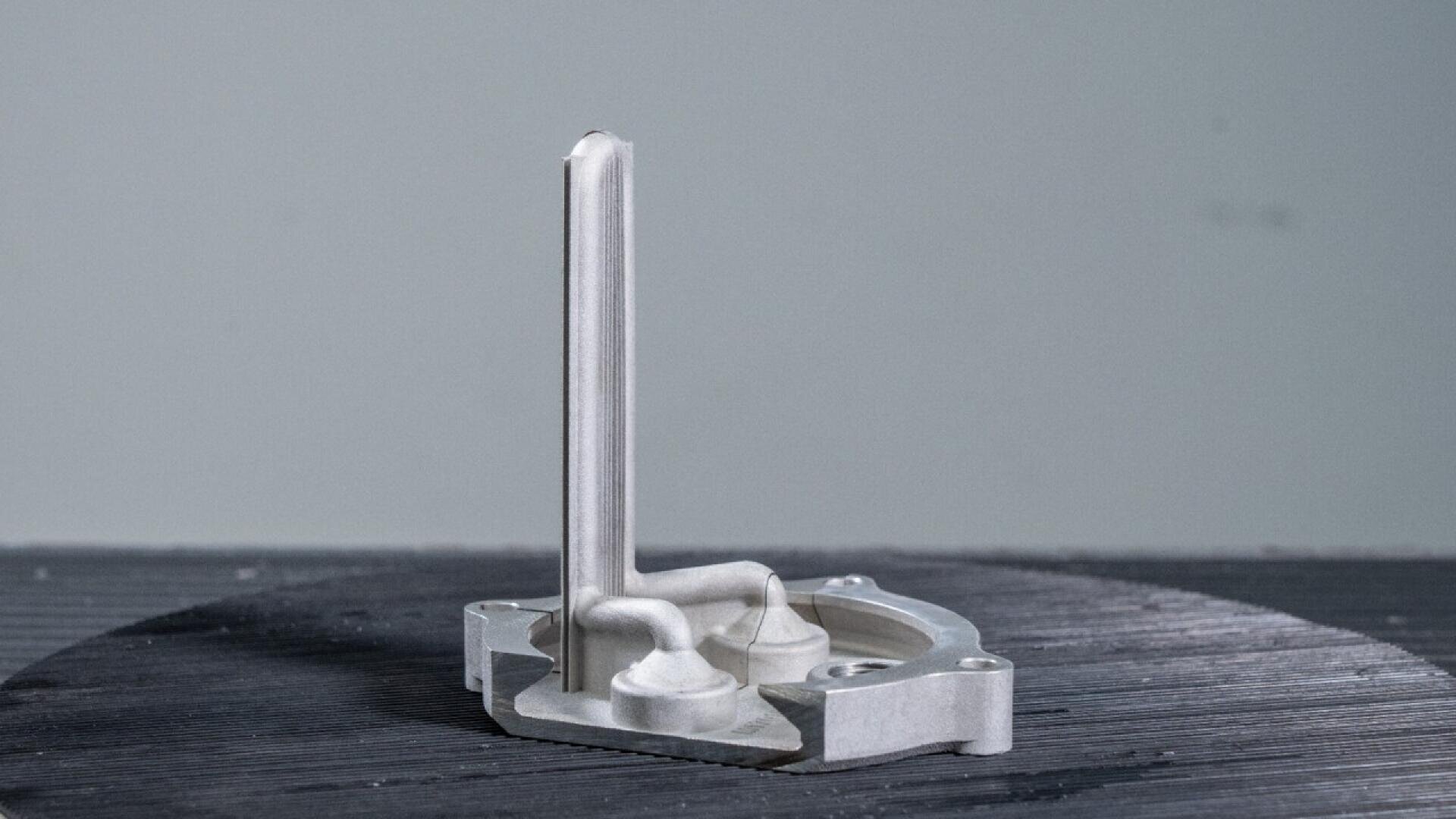 A close-up of a metallic 3D-printed industrial part with a flat circular base and two curved supports attached to a tall, vertical central structure, set against a neutral background.