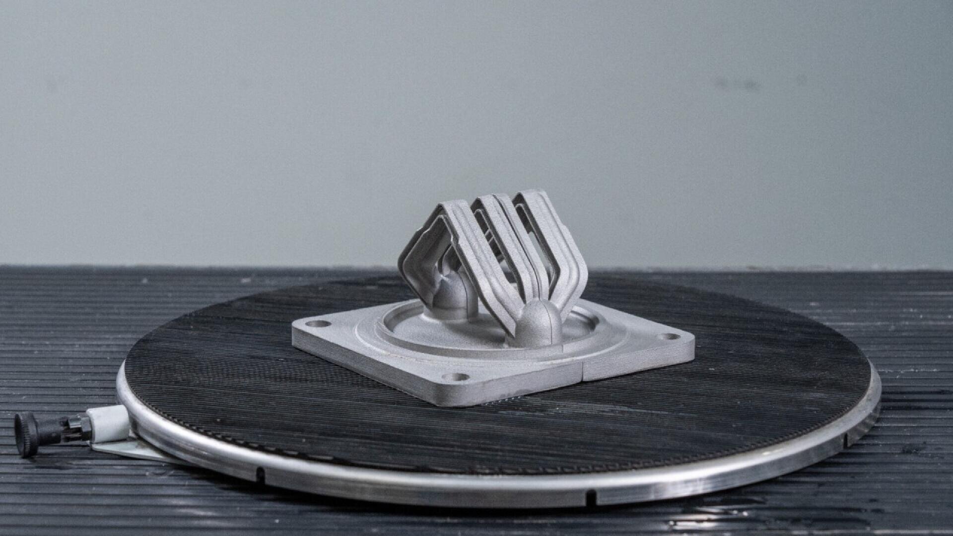 A metallic 3D-printed object with geometric angles sits on a circular platform, likely inside a manufacturing or prototyping facility. The background is neutral and uncluttered.