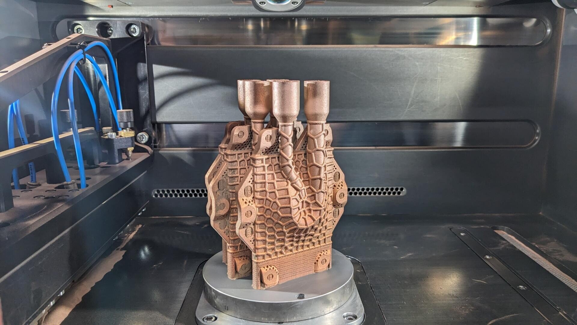 Ein 3D-Drucker druckt ein komplexes, geometrisches Metallobjekt mit Gittermustern und mehreren zylindrischen Verlängerungen, das auf einer kreisförmigen Metallplattform im Inneren des Druckers ruht. Auf der linken Seite sind blaue Schläuche sichtbar.