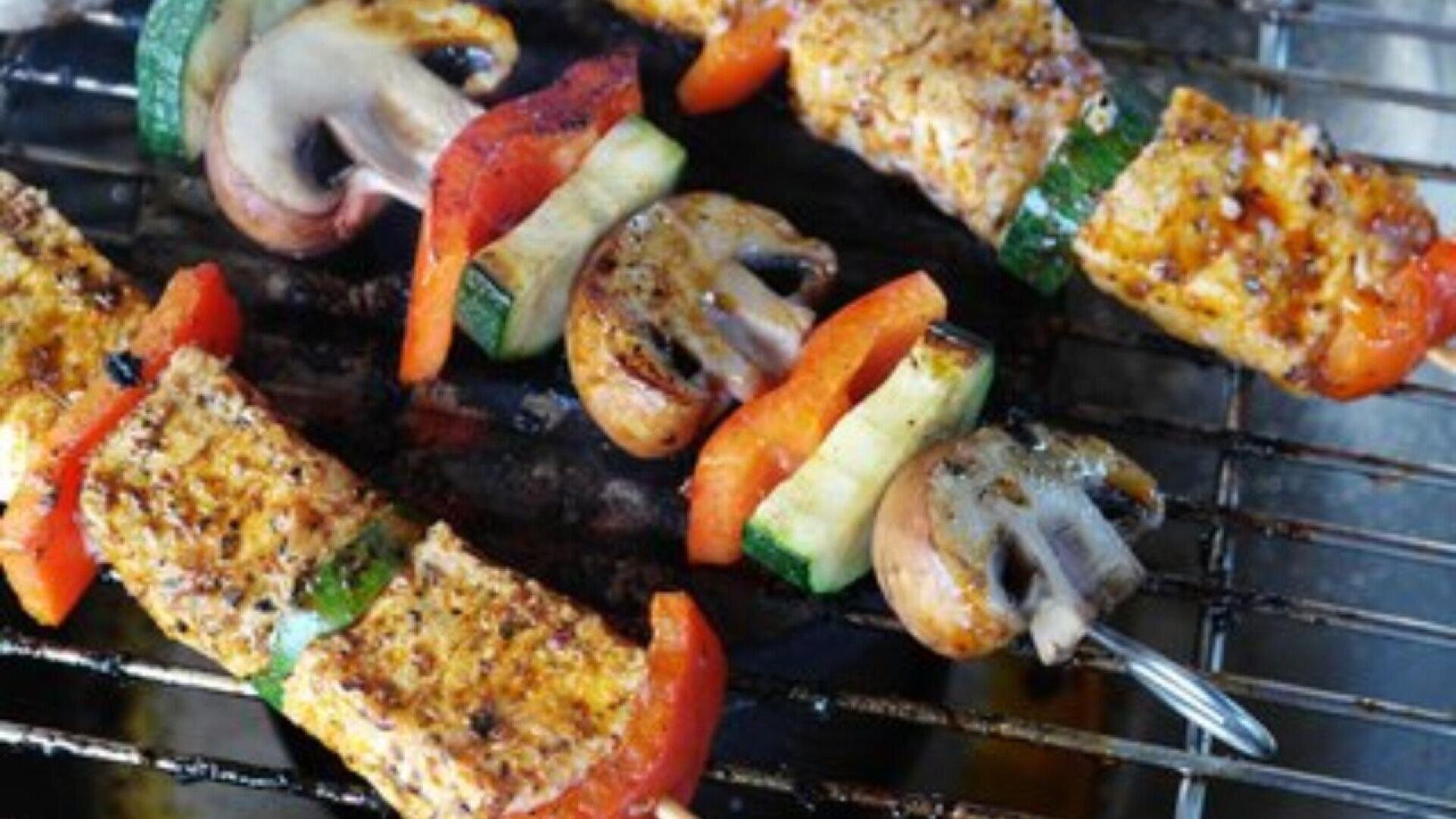 Vegetable and tofu skewers with mushrooms, zucchini, red bell peppers, and seasoned tofu pieces grilling on a barbecue.