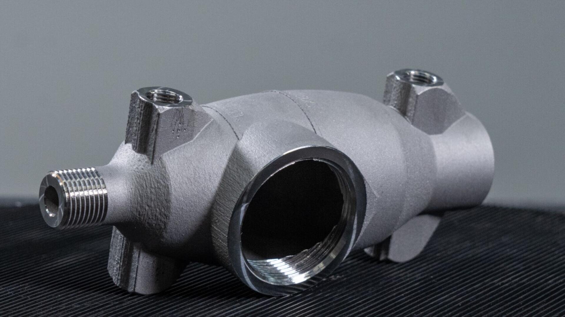A metallic, cylindrical industrial part with threaded openings on three sides and smooth surfaces, placed on a dark textured surface against a gray background.