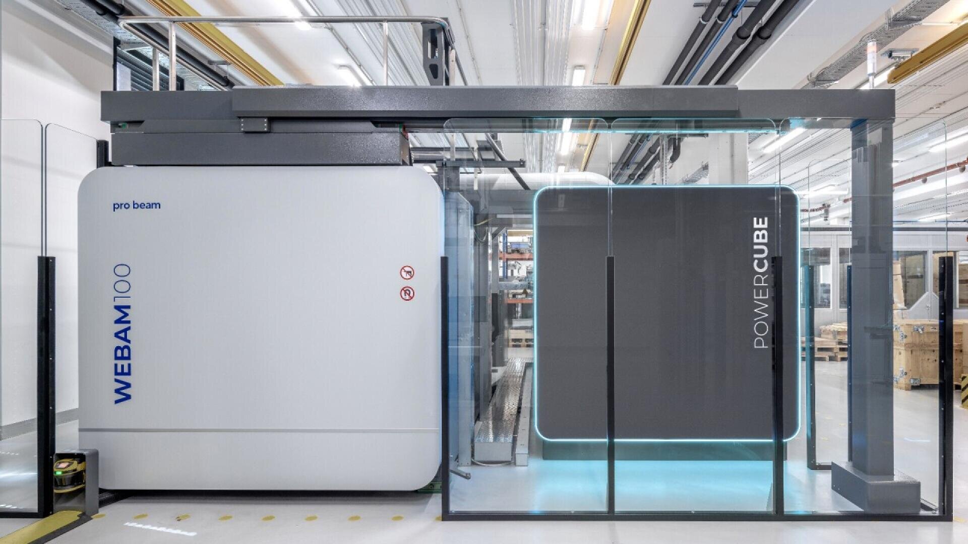 A large, modern industrial machine labeled WEBAM 100 and POWER CUBE with clean, gray and white panels, glass walls, and overhead pipes, located in a bright, well-lit facility.