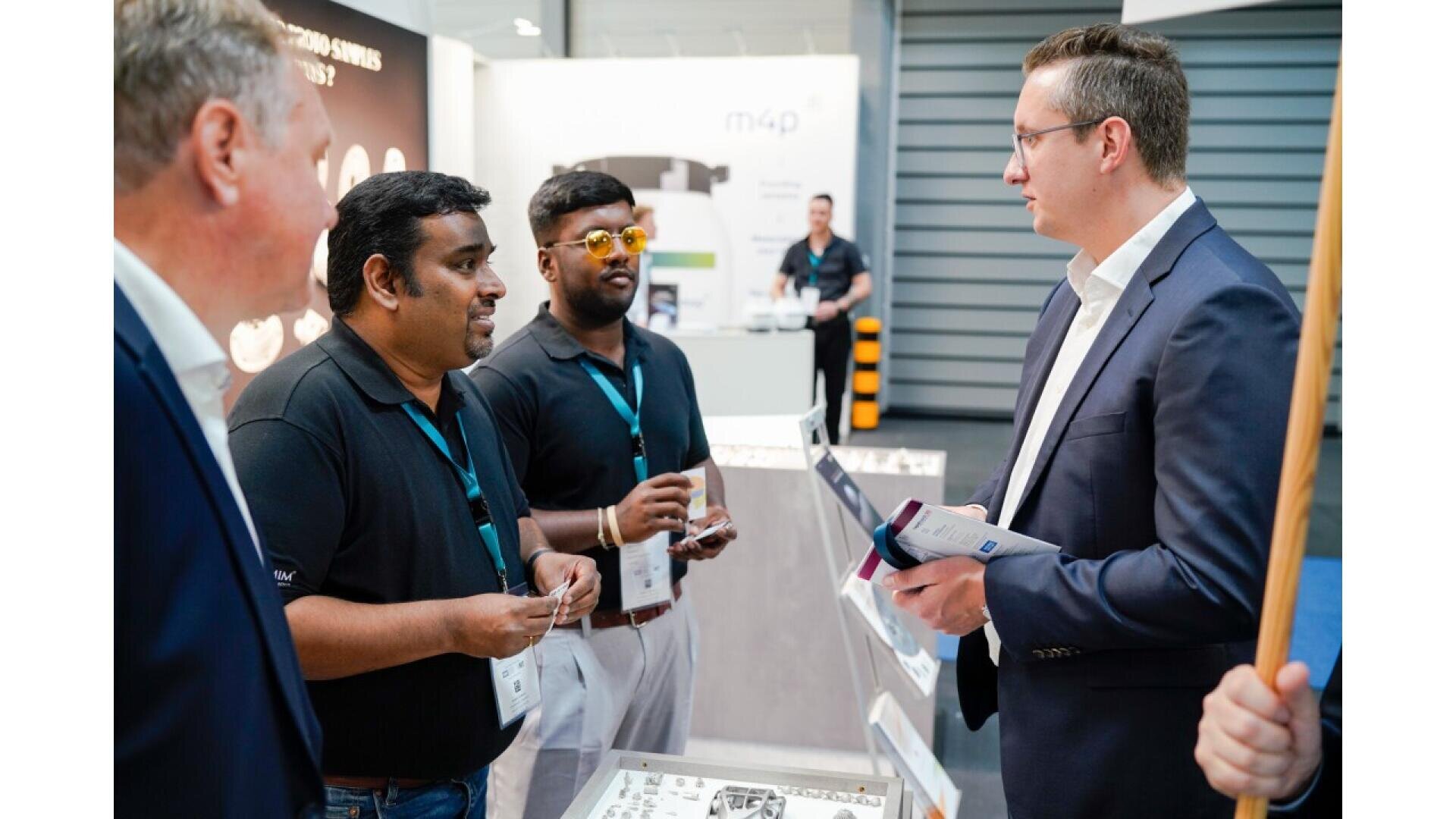 Three men in black shirts speak with a man in a suit at a trade show booth, with brochures and display items on the table between them. A fourth person stands partially visible on the left.