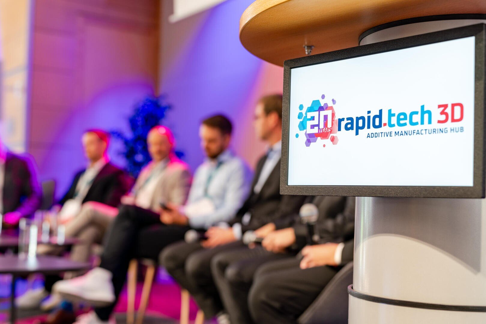 Eine Gruppe von Personen sitzt auf einer Konferenzbühne. Im Vordergrund ist ein Bildschirm mit dem Logo des rapid.tech 3D ADDITIVE MANUFACTURING HUB zu sehen. Der Hintergrund ist sanft in violetten Tönen beleuchtet.
