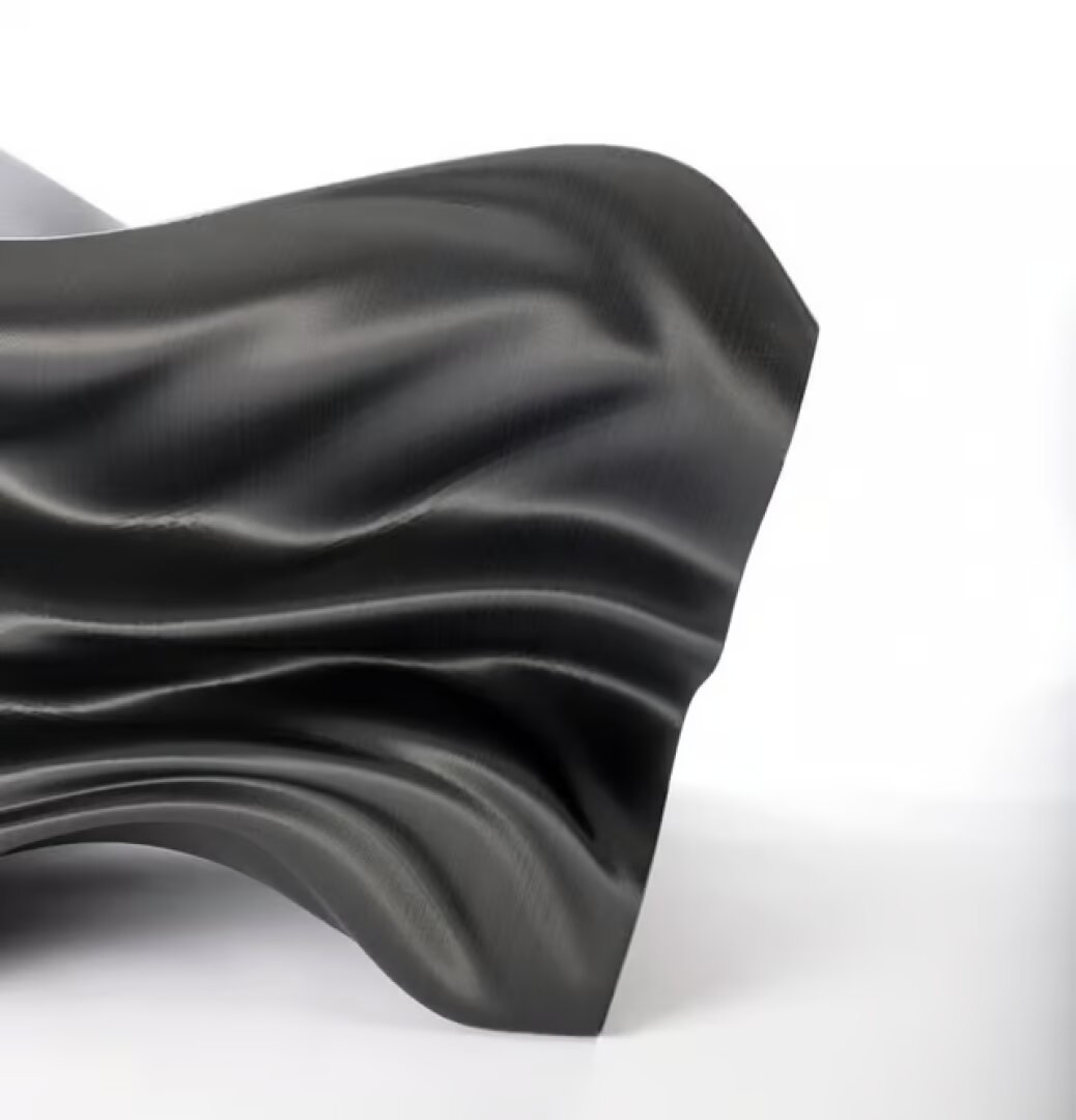 Ein glattes, schwarzes, flexibles Blatt mit wellenförmigen, fließenden Falten wird vor einem weißen Hintergrund angezeigt, wodurch ein abstrakter und skulpturaler Effekt entsteht.