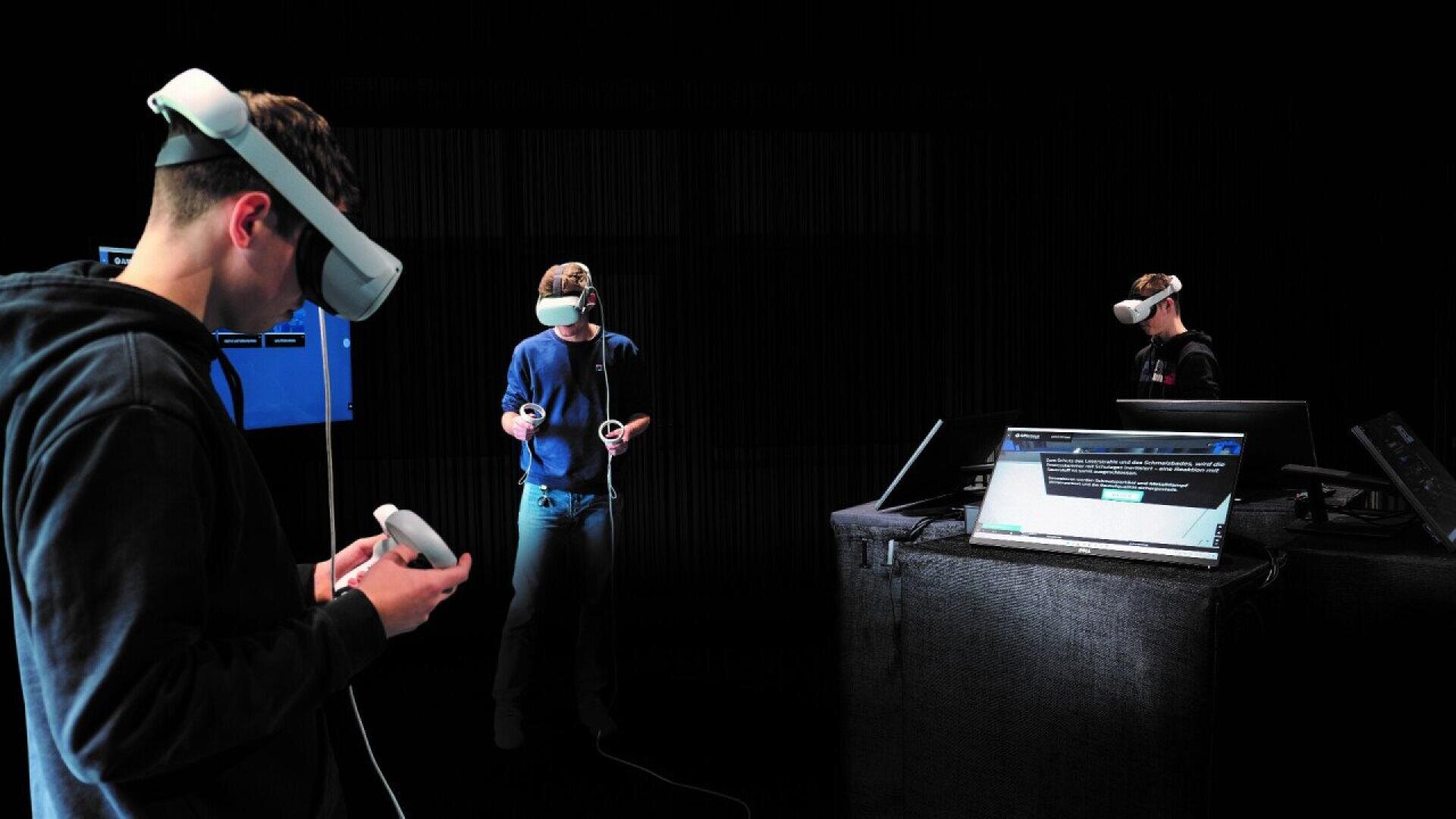 Three people wearing VR headsets and holding controllers stand in a dark room with computers and screens nearby, interacting with virtual content.