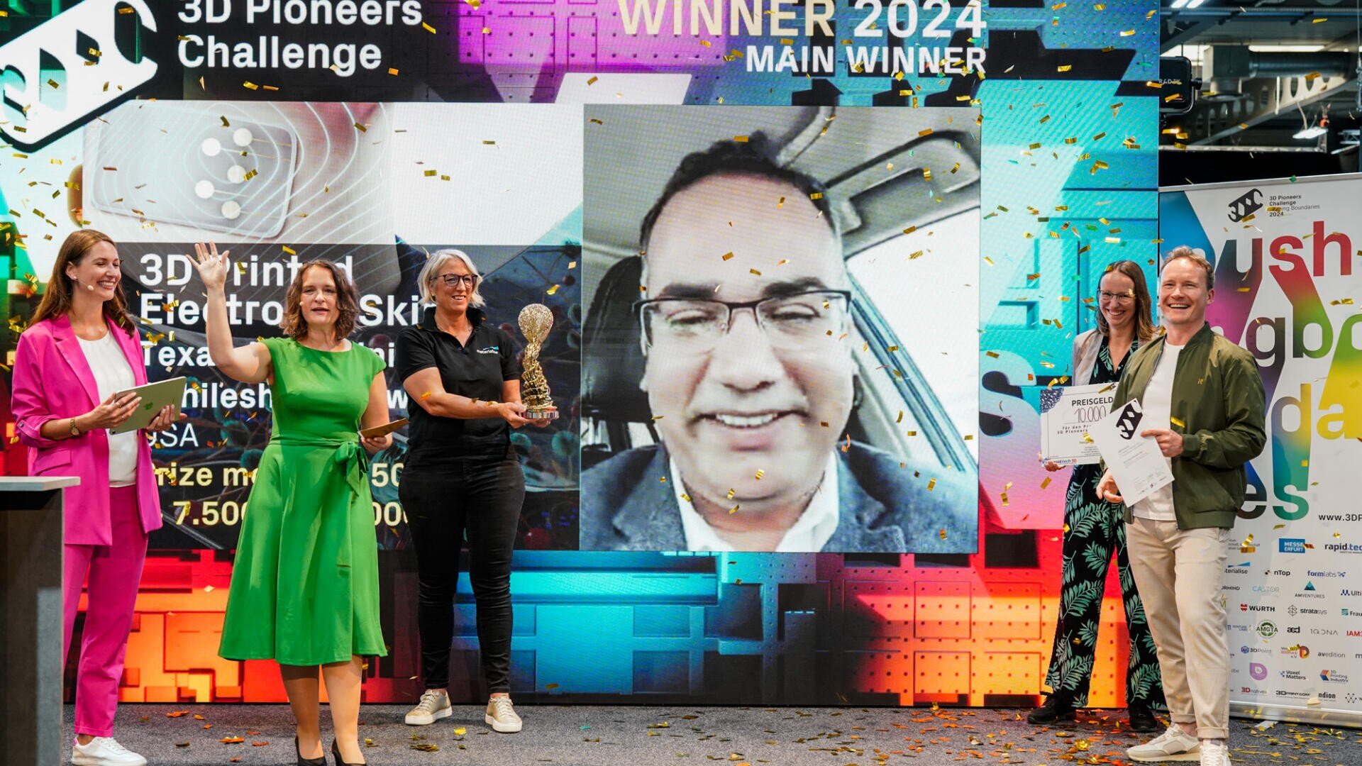 Eine Gruppe von Menschen feiert auf der Bühne der 3D Pioneers Challenge 2024 mit Trophäe und Urkunde, während auf einer großen Videoleinwand hinter ihnen ein lächelnder Mann erscheint. Konfetti regnet, während sie für ein Foto posieren.