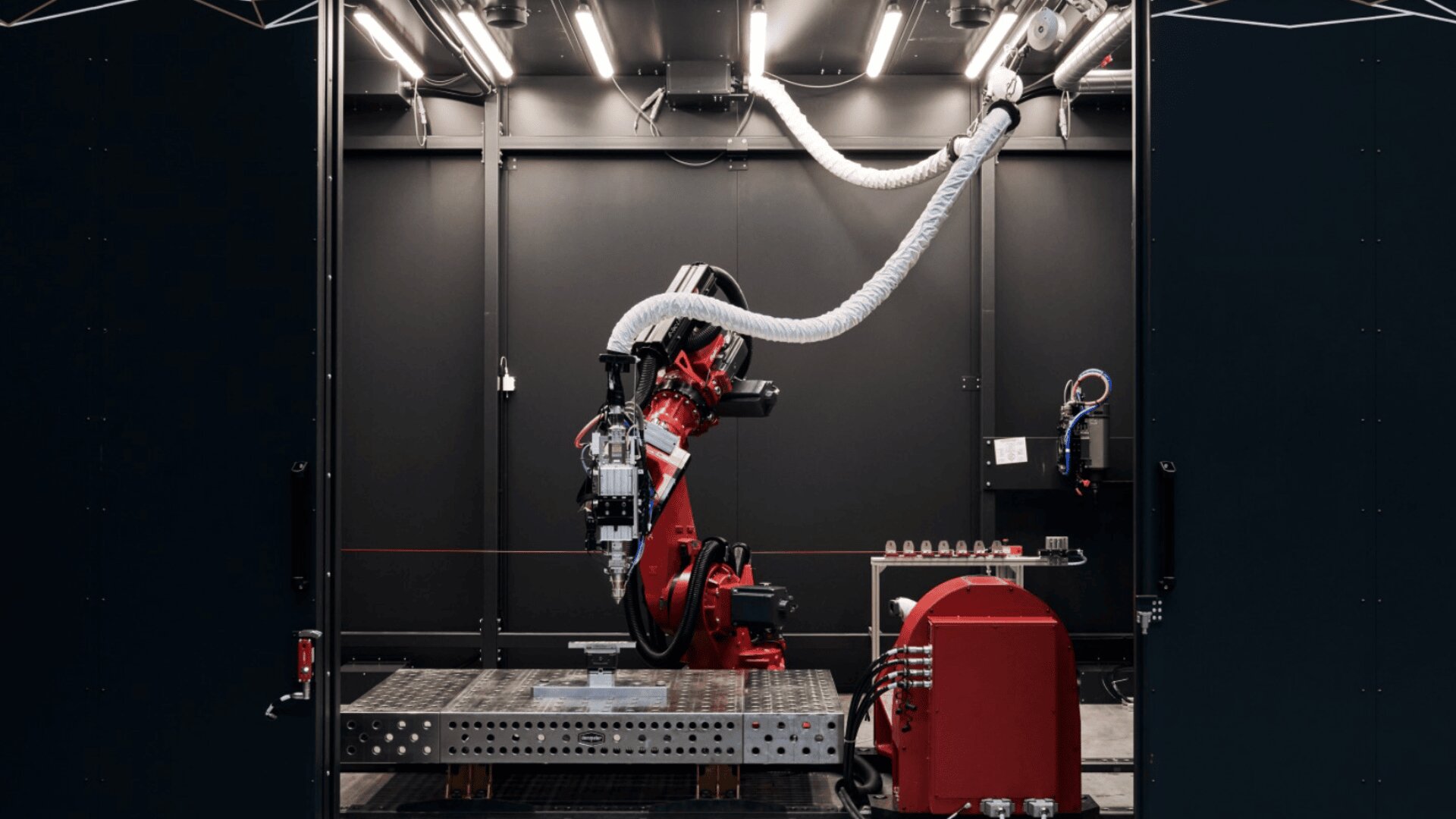 A red industrial robotic arm operates inside a dark, modern workspace, surrounded by cables and machinery, under bright overhead lights.