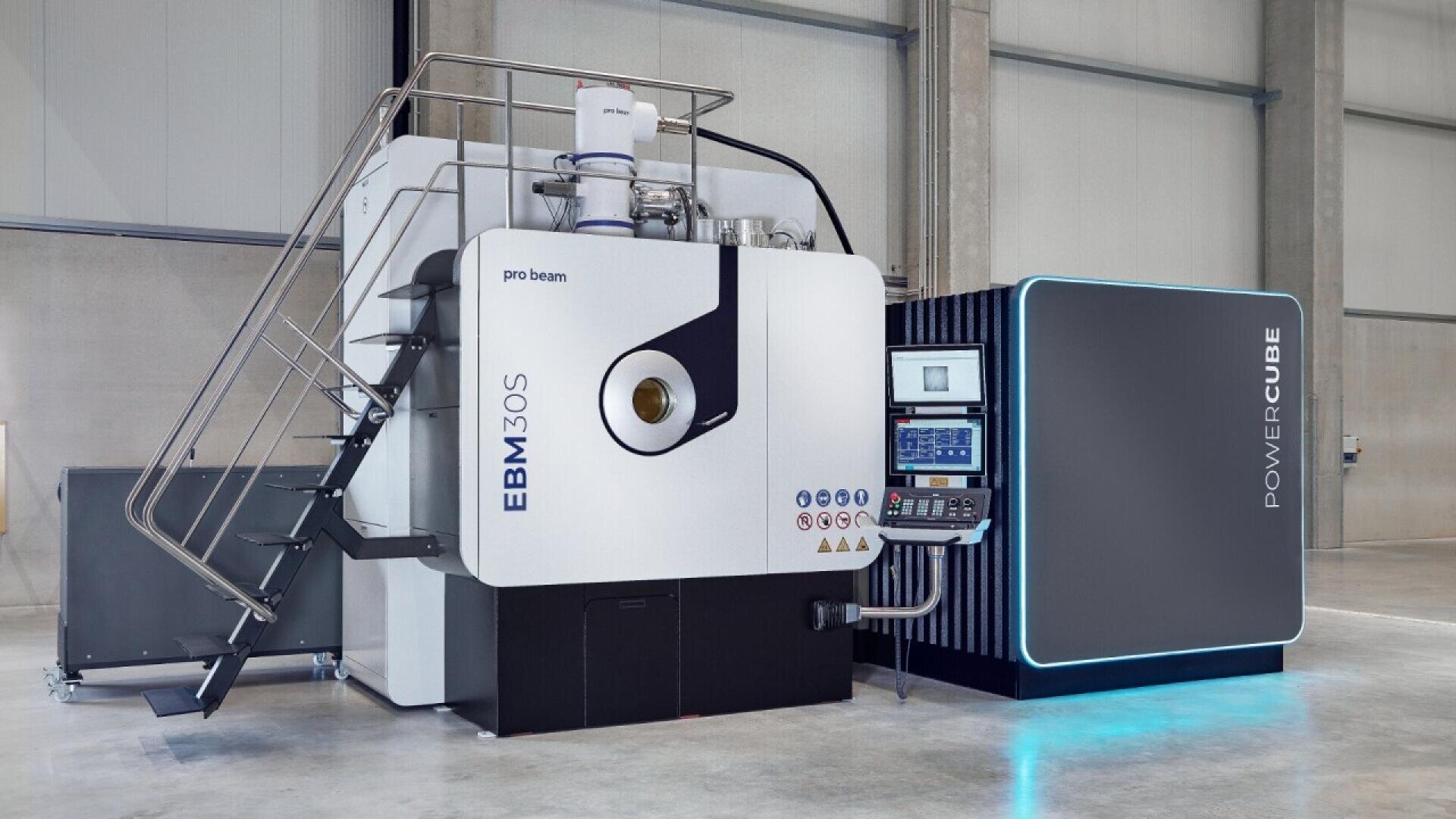 A large industrial EBM 30S machine labeled “pro beam” and “POWERCUBE” stands in a spacious, modern facility. It features control panels, a staircase, and metal pipes, used for advanced manufacturing processes.