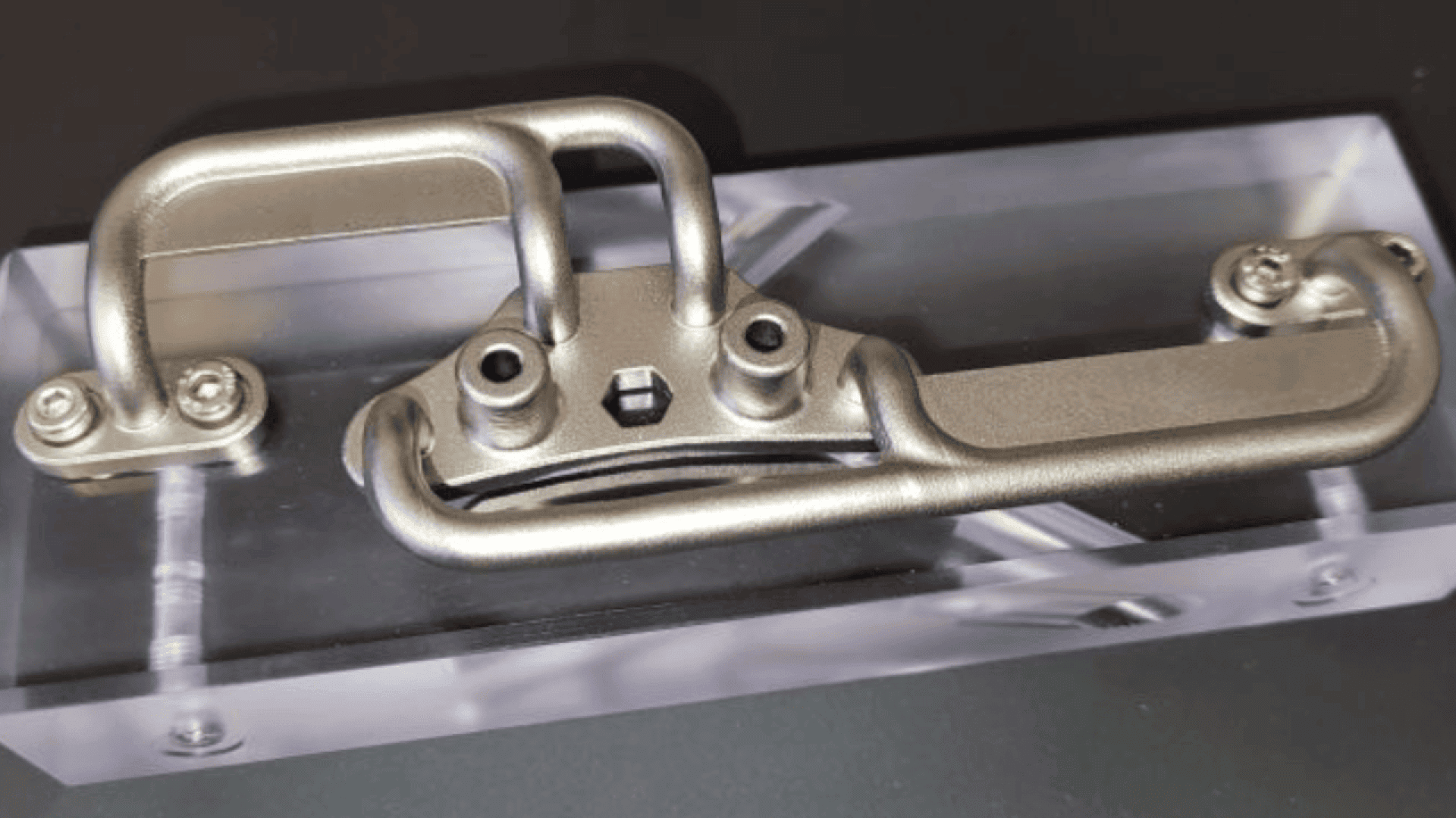 Ein komplexes Metallteil mit gebogenen und geraden Abschnitten ist auf einer durchsichtigen rechteckigen Acrylbasis montiert und an den Ecken mit Schrauben befestigt. Das Ganze ist vor einem dunklen Hintergrund angeordnet.
