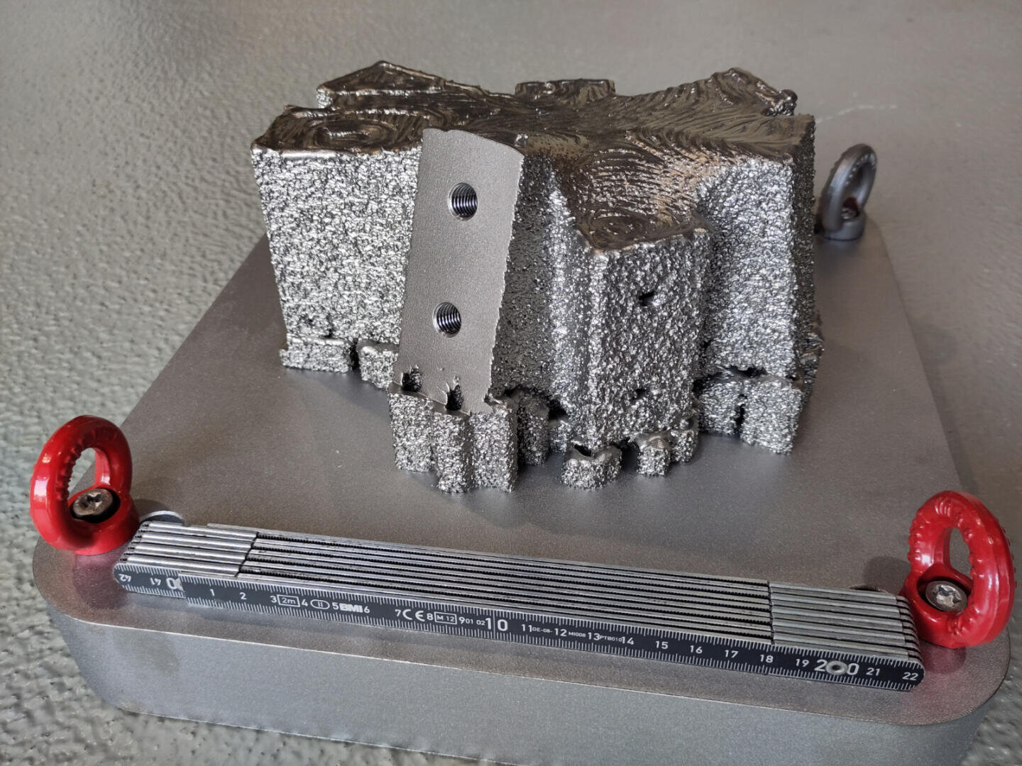 Ein grob strukturiertes, metallisches 3D-gedrucktes Teil mit zwei Löchern sitzt auf einer glatten Metallgrundplatte. Die Basis ist mit roten Hebeösen und einem Lineal mit der Aufschrift „200“ versehen.