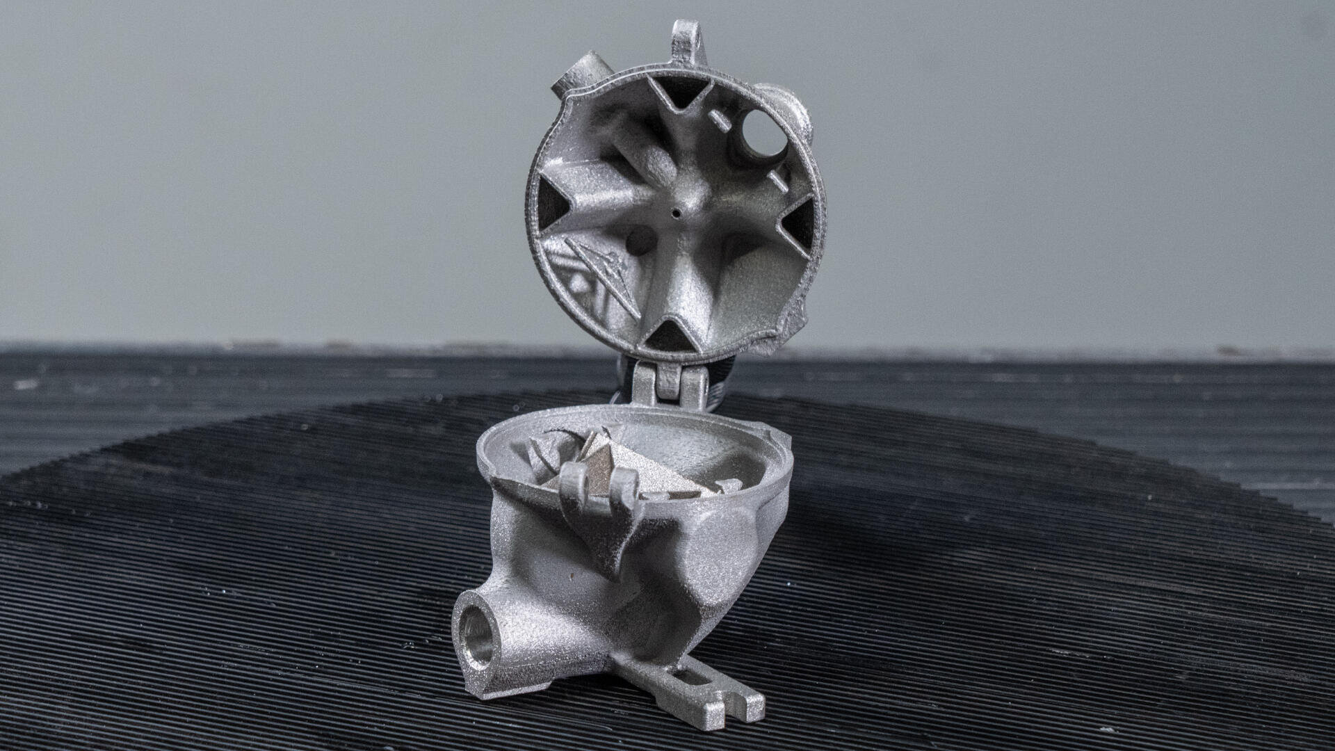 Eine silberne, industrielle Metallkomponente mit rundem Design und offenem Deckel wird aufrecht auf einer dunklen, strukturierten Oberfläche vor einem schlichten grauen Hintergrund präsentiert.