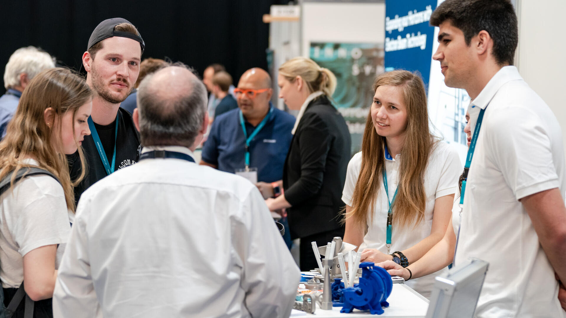 Eine Gruppe junger Erwachsener, zwei Frauen und zwei Männer, unterhält sich mit einem älteren Mann an einem Messestand, auf dem Industrieausrüstung ausgestellt ist. Alle tragen Namensschilder und scheinen in ein Gespräch vertieft zu sein.