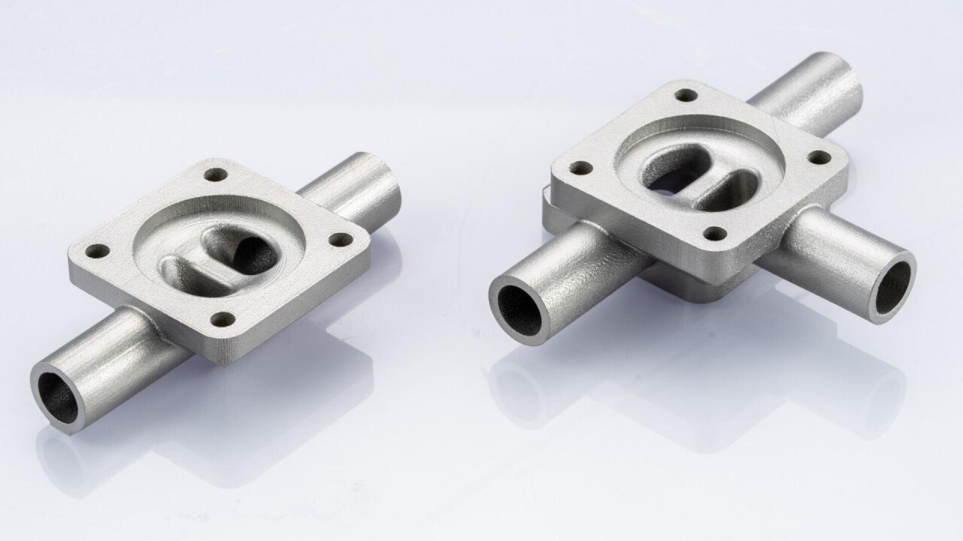 Two metallic machined components with rectangular bases, four bolt holes at the corners, central cavities, and cylindrical tubes extending from opposite sides, placed on a white surface.