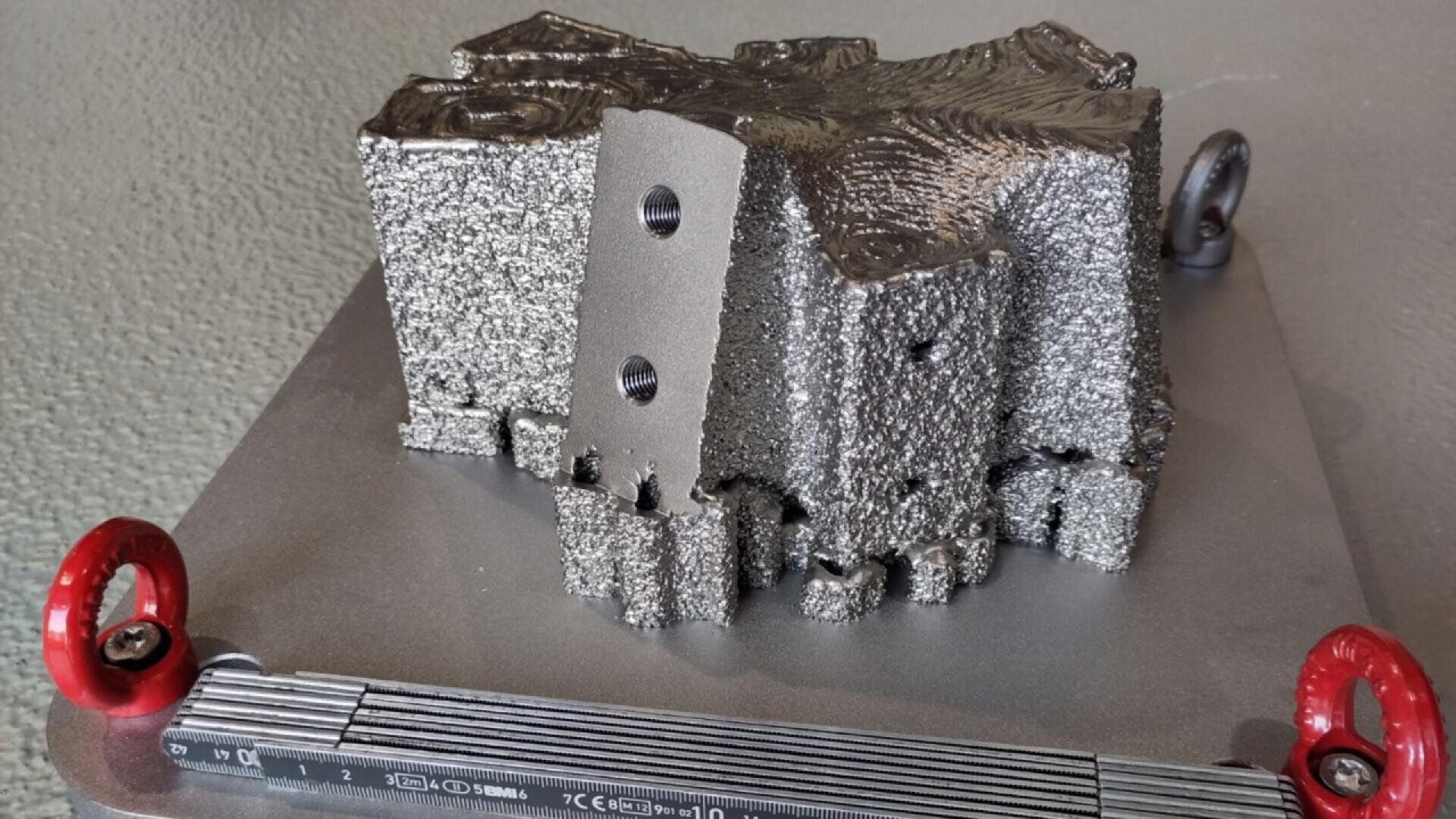 A metallic 3D-printed object with a rough, textured surface and drilled holes sits on a flat metal base with red ring handles at the corners. The base has measurement markings on one side.