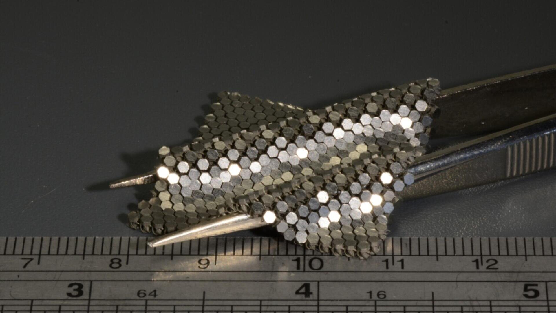 A close-up of metallic, hexagon-patterned objects held by tweezers, with a ruler showing centimeter and millimeter measurements below them. The objects resemble small wing shapes.
