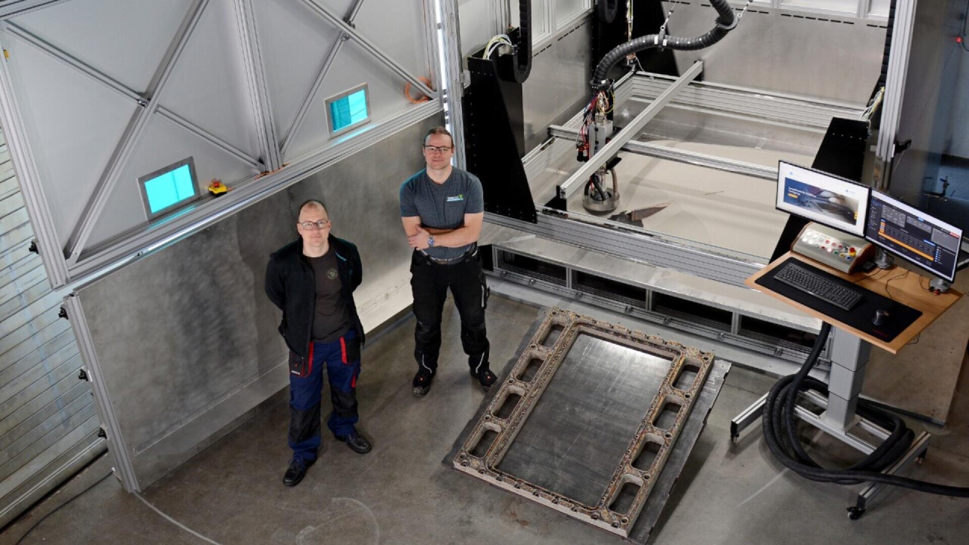 Two people stand in an industrial workspace next to a large piece of machinery, a metal frame on the floor, and a workstation with two computer monitors displaying technical data.