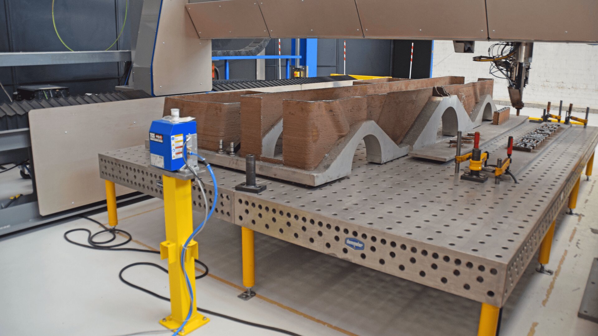 Ein großer 3D-Drucker erstellt eine Struktur mit bogenförmigen Formen aus braunem Material auf einem Arbeitstisch aus Metall, und um den Arbeitsbereich herum sind Klemmen und Geräte zu sehen.