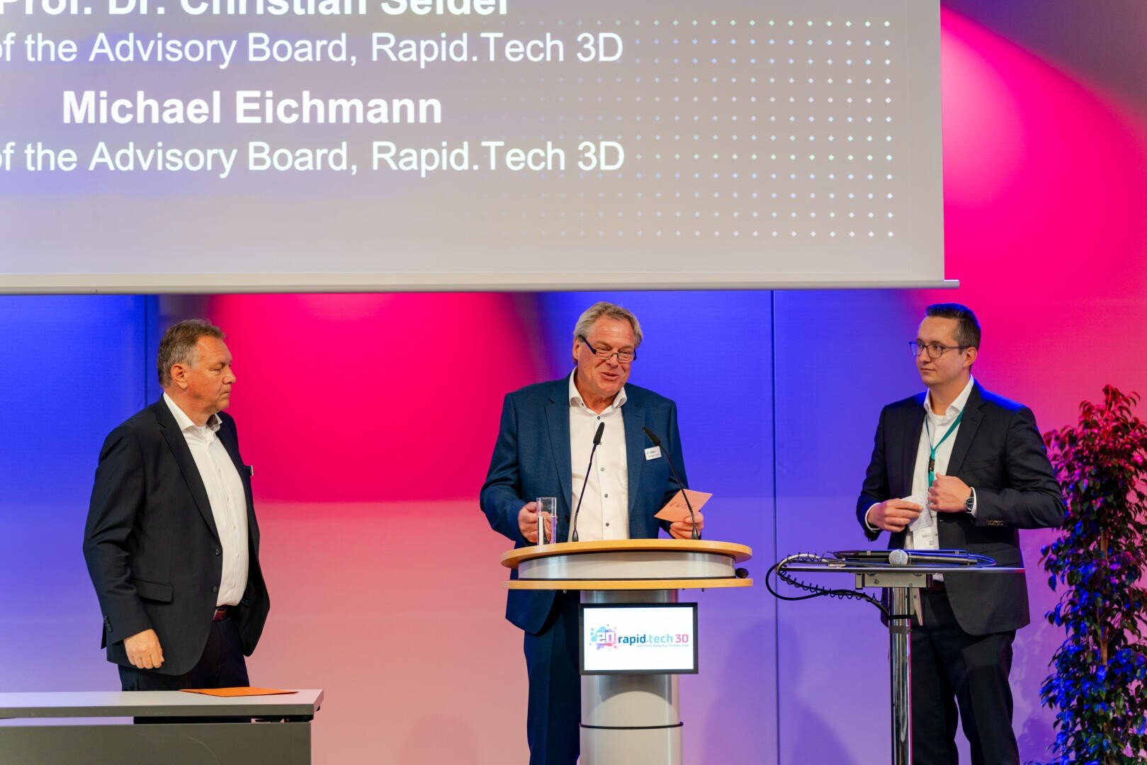 Drei Männer in Anzügen stehen während einer Präsentation auf einer Bühne. Einer spricht an einem Podium, die anderen hören zu. Hinter ihnen ist eine Leinwand mit den Namen Prof. Dr. Christian Seider und Michael Eichmann zu sehen.