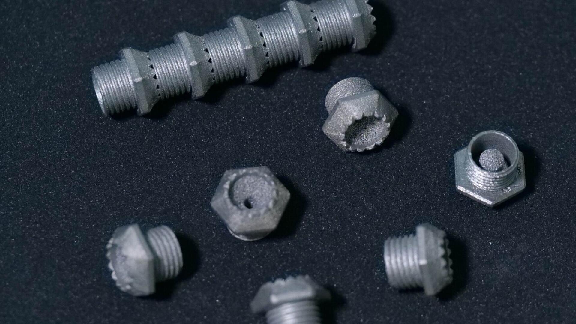 Several metallic, hexagonal bolts and a single longer threaded rod with textured surfaces are arranged on a black surface. The objects have a rough, matte finish and distinct threading details.
