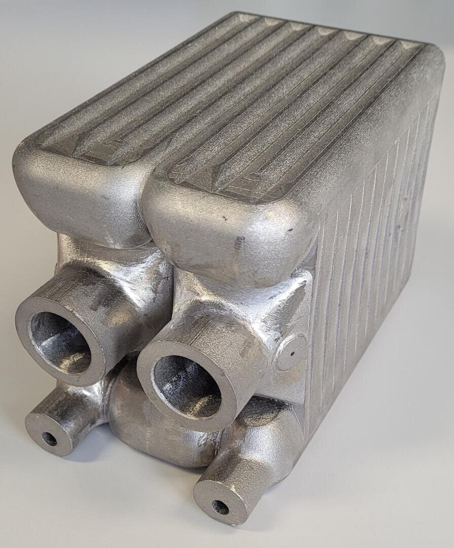 A metallic, rectangular heat exchanger with four cylindrical pipe connections and a ribbed surface, placed on a light gray background.
