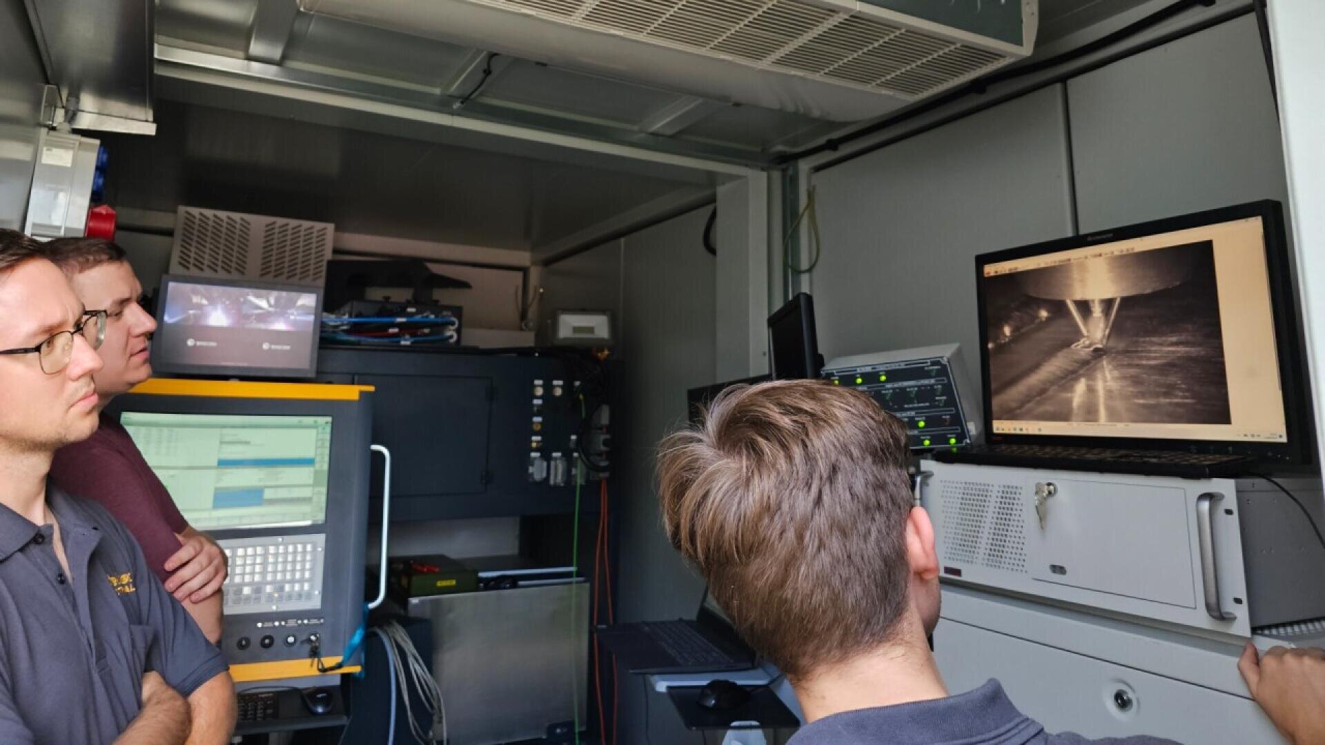 Three people in a control room look at a monitor displaying technical or scientific data. One person is pointing at the screen, and equipment and computers are visible in the background.