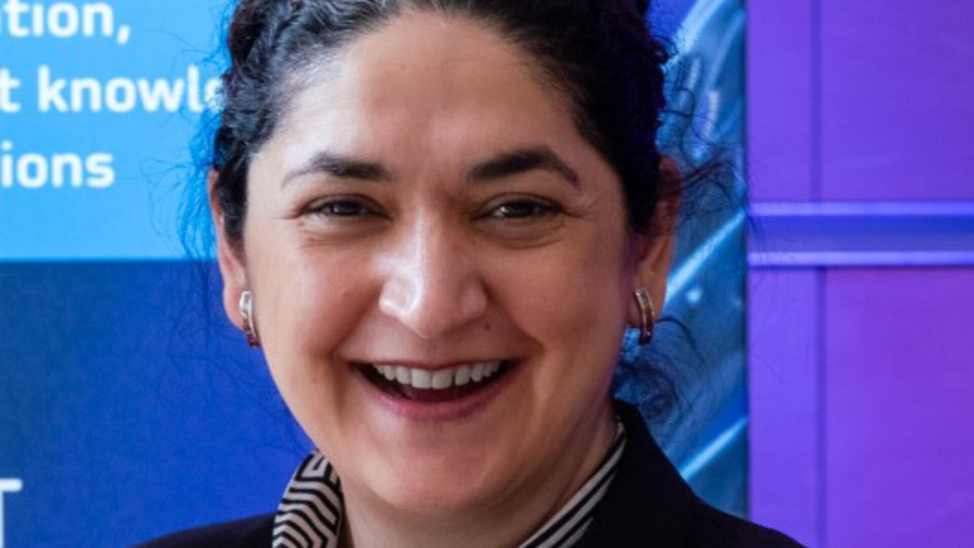 Eine Frau mit zurückgekämmtem dunklem, lockigem Haar, einem schwarzen Blazer und einem gestreiften Hemd lächelt in die Kamera. Der Hintergrund ist blau und lila mit teilweise sichtbarem weißem Text.