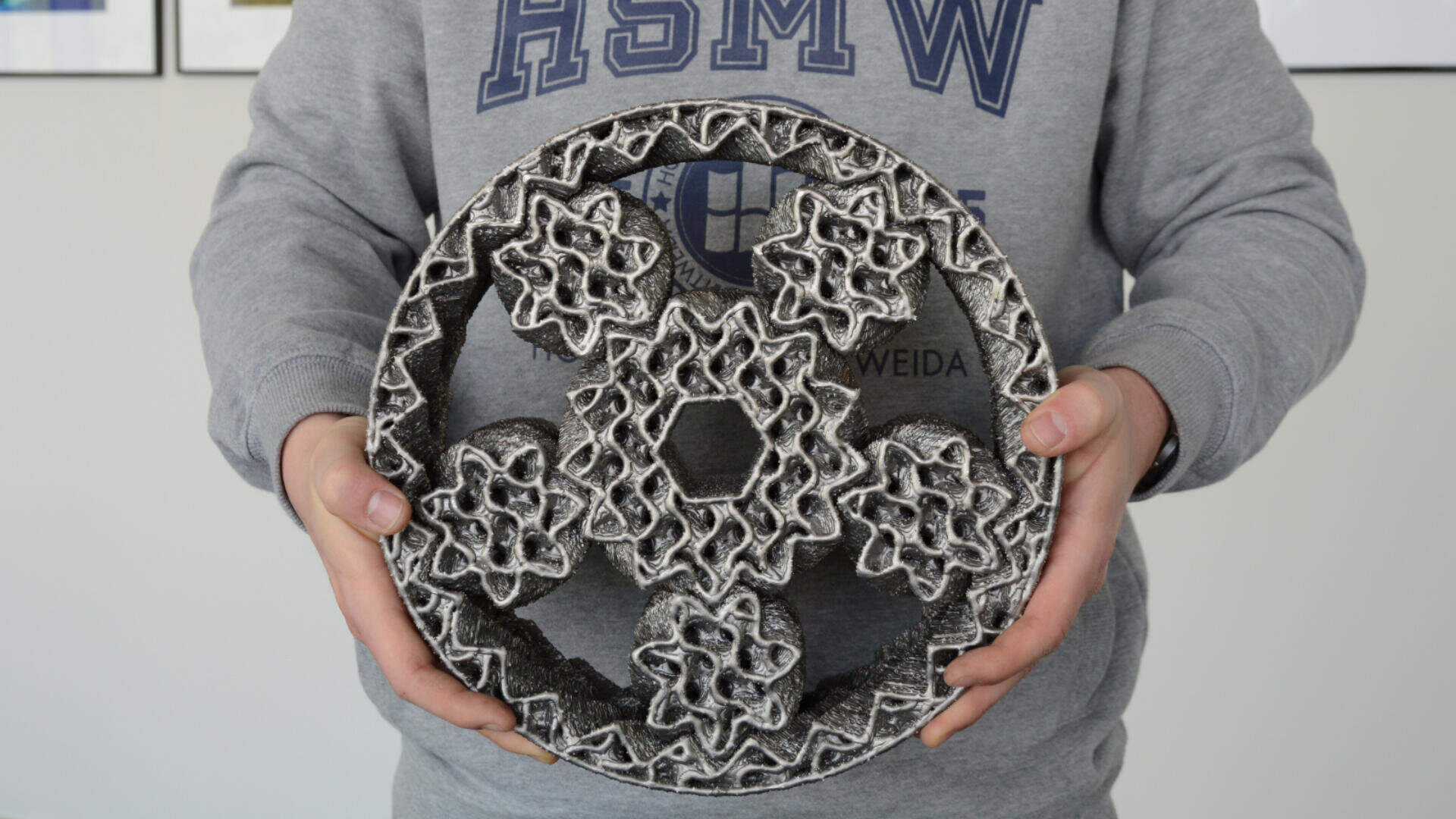 Eine Person in einem grauen Sweatshirt hält ein großes, kompliziertes, kreisförmiges 3D-gedrucktes Objekt mit einem geometrischen, gitterartigen Design und einer metallischen Textur.