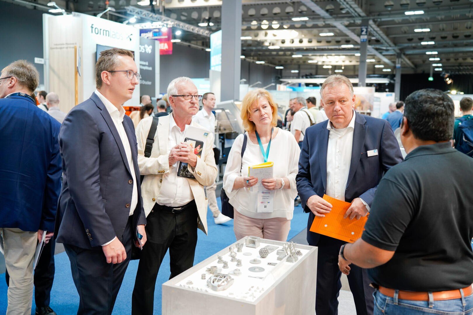 Eine Gruppe professionell gekleideter Menschen steht auf einer Ausstellung oder Handelsmesse um einen Ausstellungstisch mit Metallgegenständen herum und hört einer Person aufmerksam zu, die mit ihnen spricht.