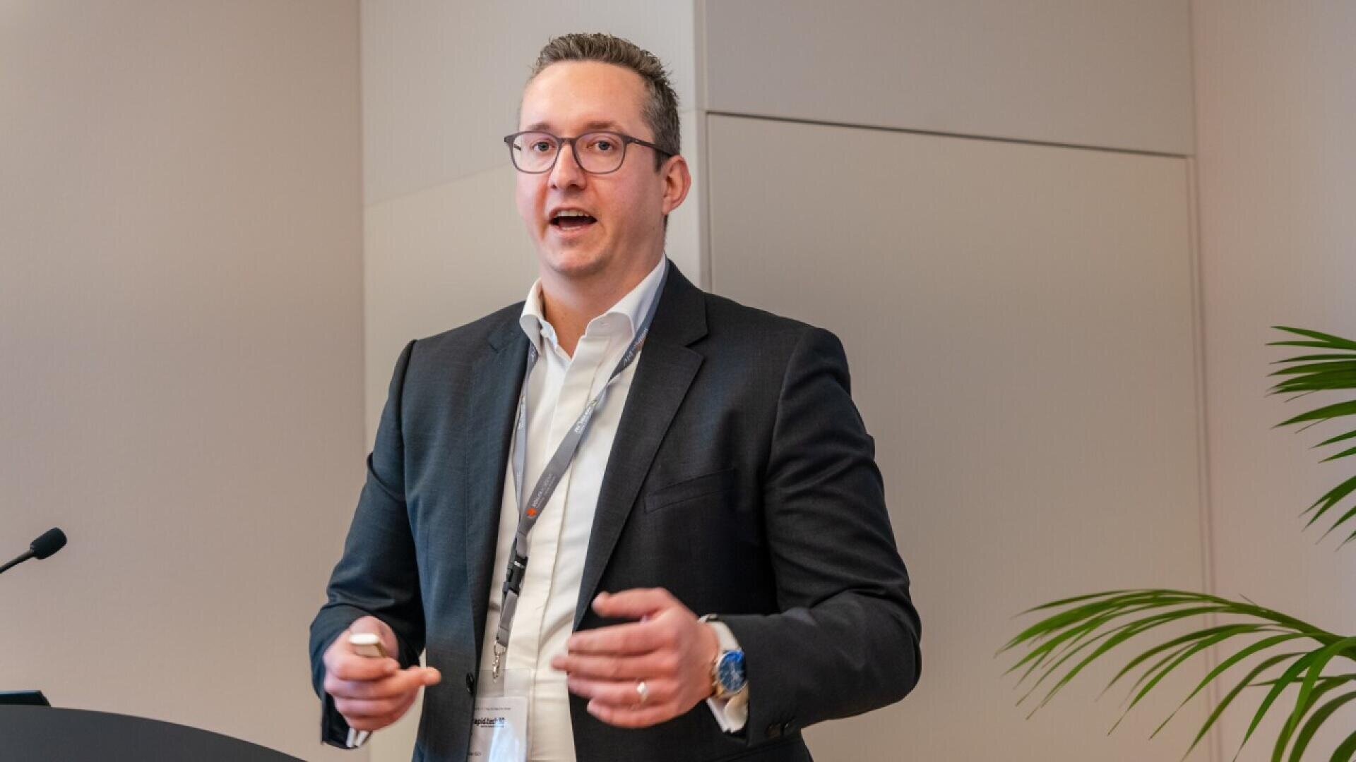 A man in a dark suit and glasses gives a presentation in an office setting, speaking and gesturing with his hands. A microphone and a plant are visible in the foreground.