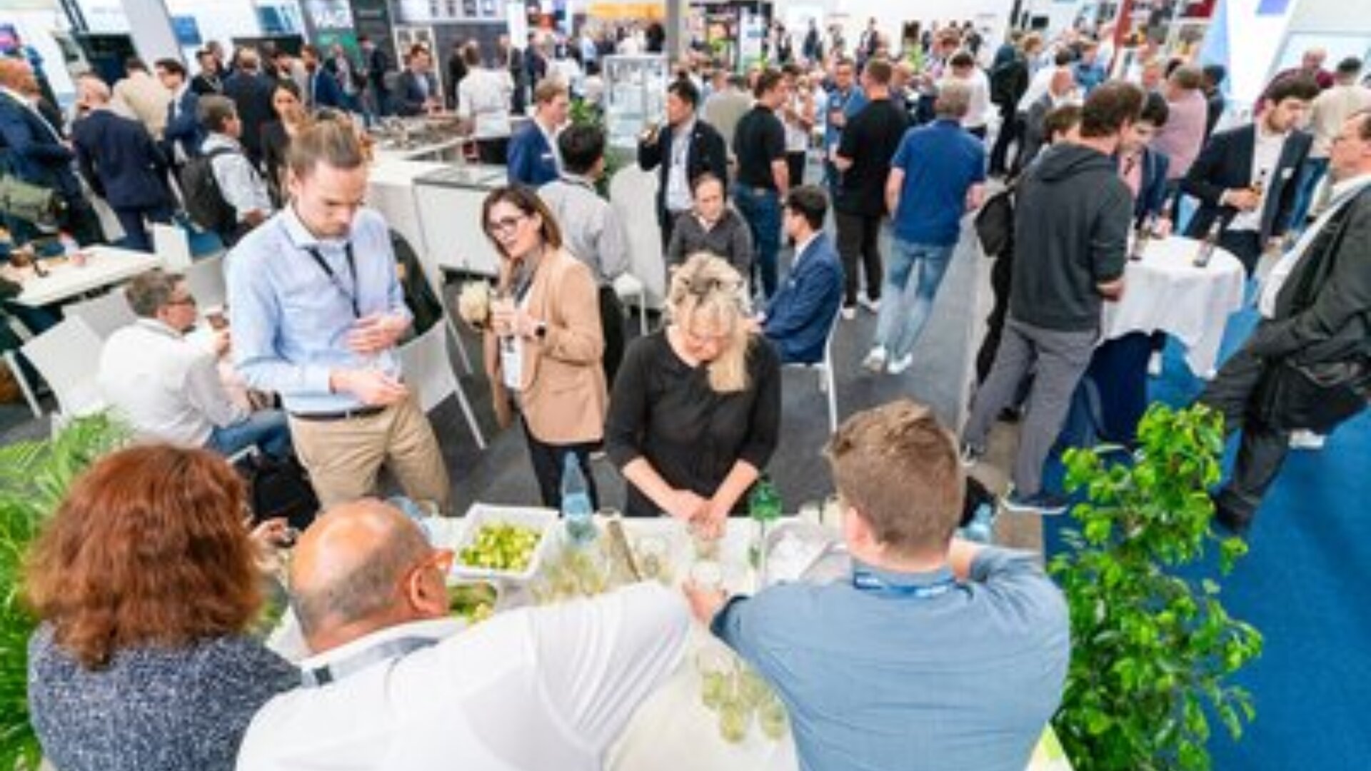 Eine große Gruppe von Menschen trifft sich bei einem geschäftigen Networking-Event in einer Halle. Einige stehen, andere sitzen an Tischen mit Getränken und Snacks in einem hellen, modernen Raum.