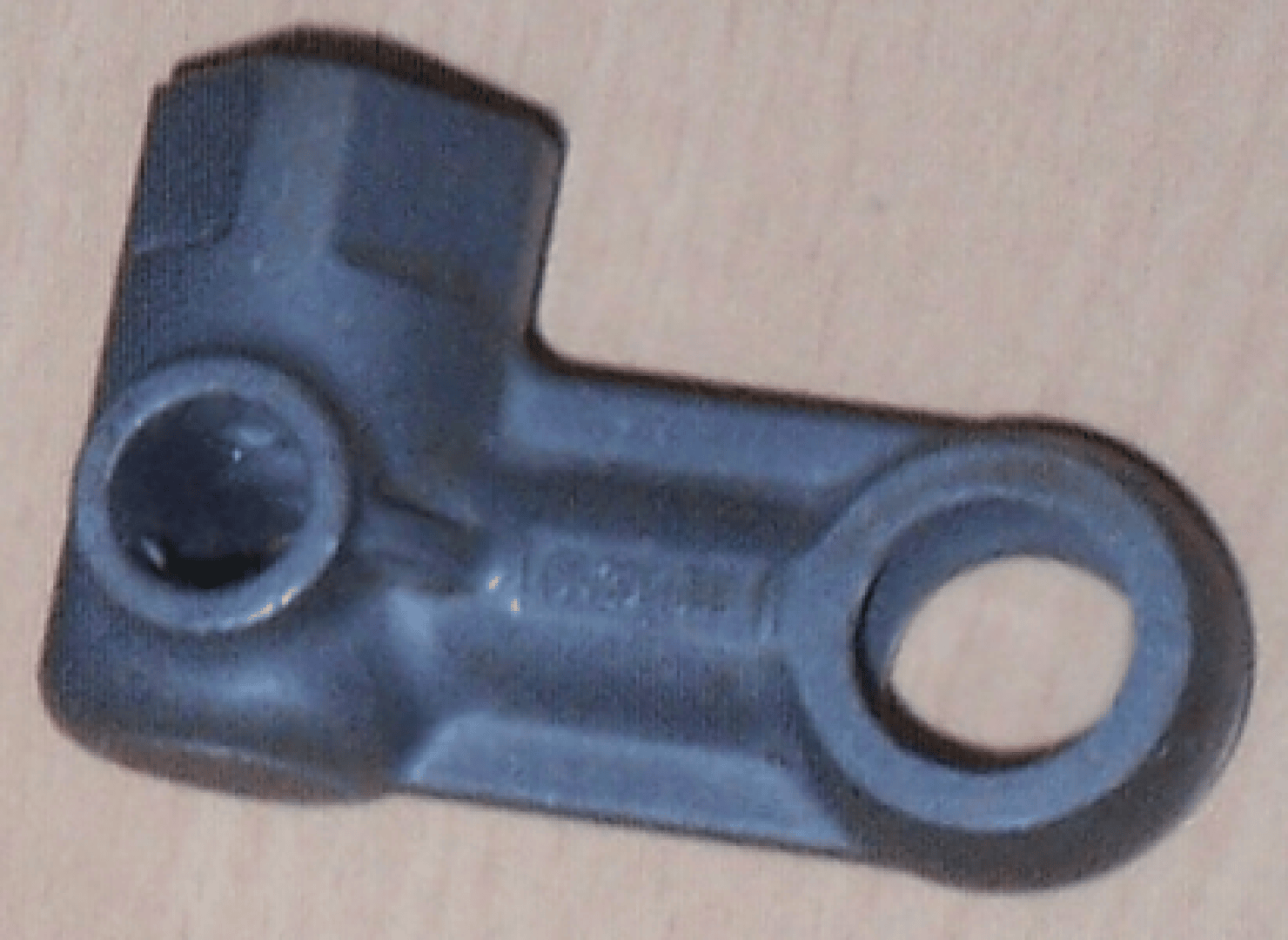 Ein schwarzes, L-förmiges Kunststoff- oder Metallteil mit einer runden und einer sechseckigen Öffnung, das auf einer hellen Holzoberfläche liegt.
