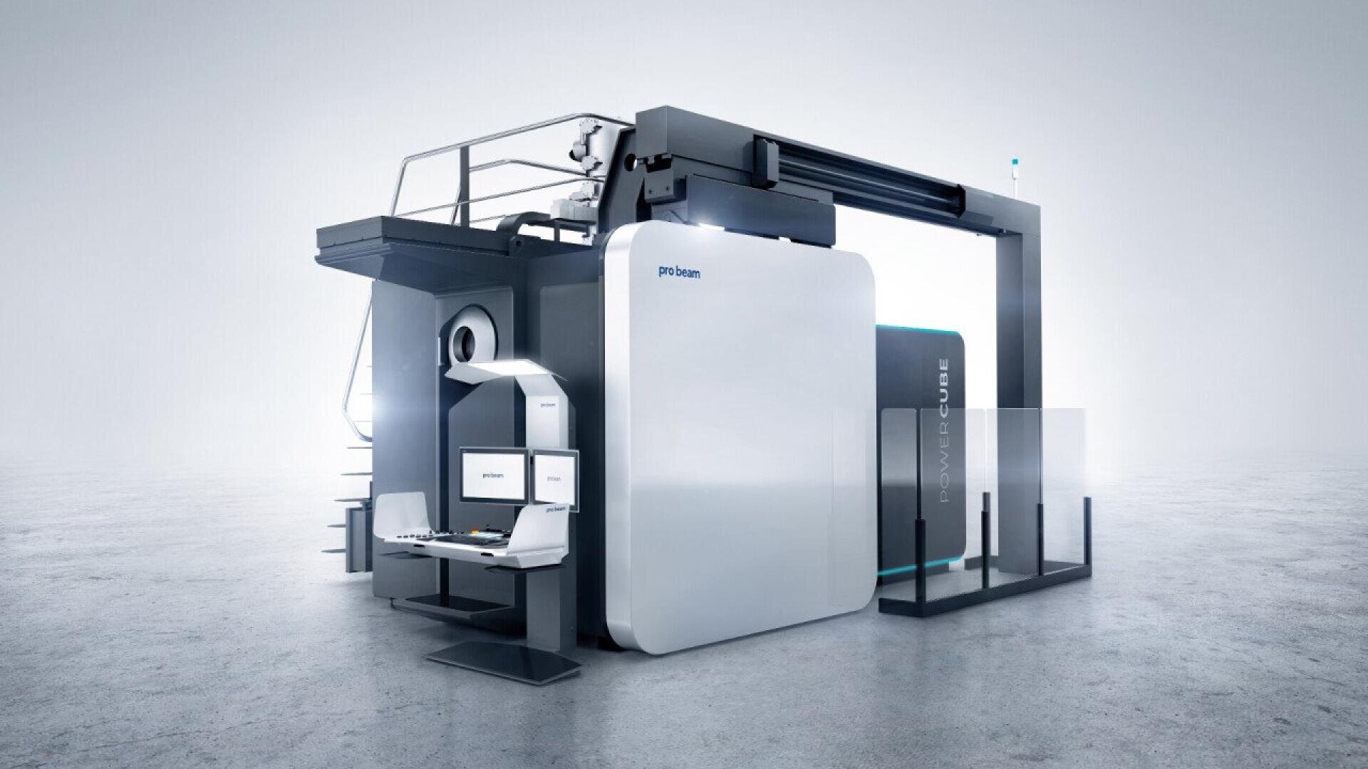 A large, modern industrial 3D printer with a sleek white and gray exterior, dual computer workstations, and an elevated platform, set in a spacious, well-lit environment.