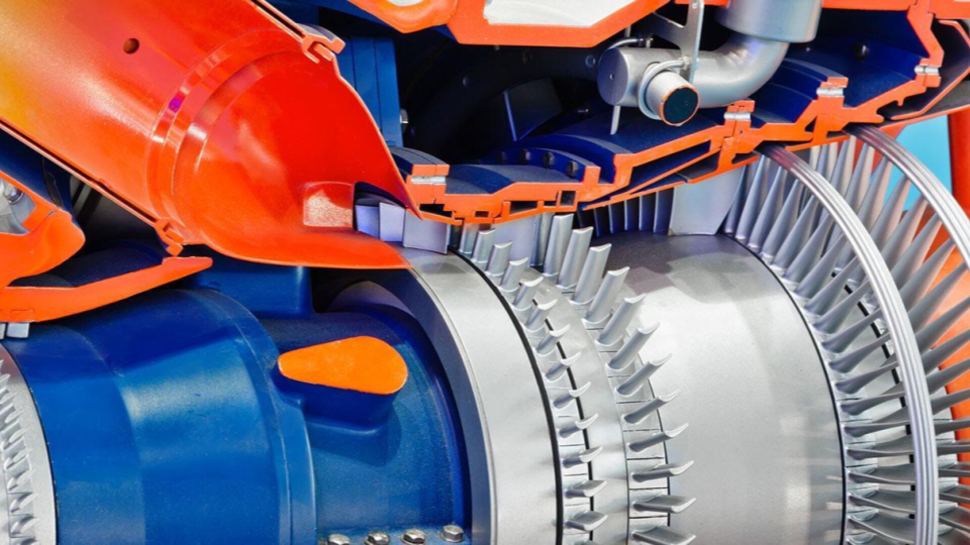 Nahaufnahme des Querschnitts eines Düsentriebwerks mit silberfarbenen Turbinenschaufeln, blauen und orangefarbenen Komponenten und verschiedenen mechanischen Teilen im Inneren des Triebwerks.