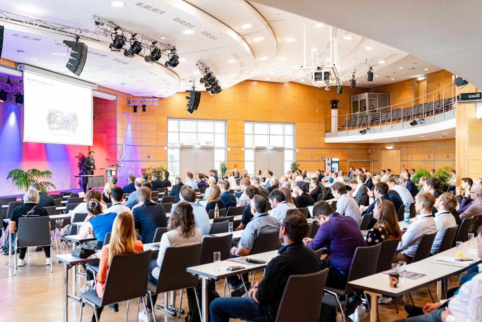 Eine große Gruppe von Menschen sitzt an Tischen in einem modernen Konferenzsaal. Vor ihnen steht eine Bühne mit einem Moderator und einer großen Leinwand, auf der eine Präsentation läuft. Der Raum ist hell erleuchtet, mit Holzvertäfelung und hohen Decken.