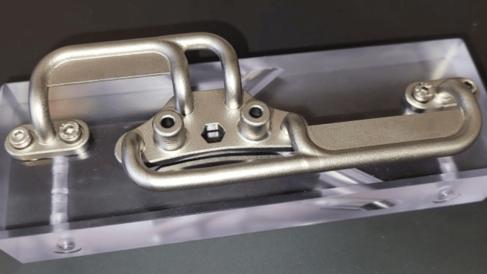 A metallic, industrial component with curved and angled tubing is mounted on a clear rectangular acrylic base, secured by four screws at the corners, against a dark background.