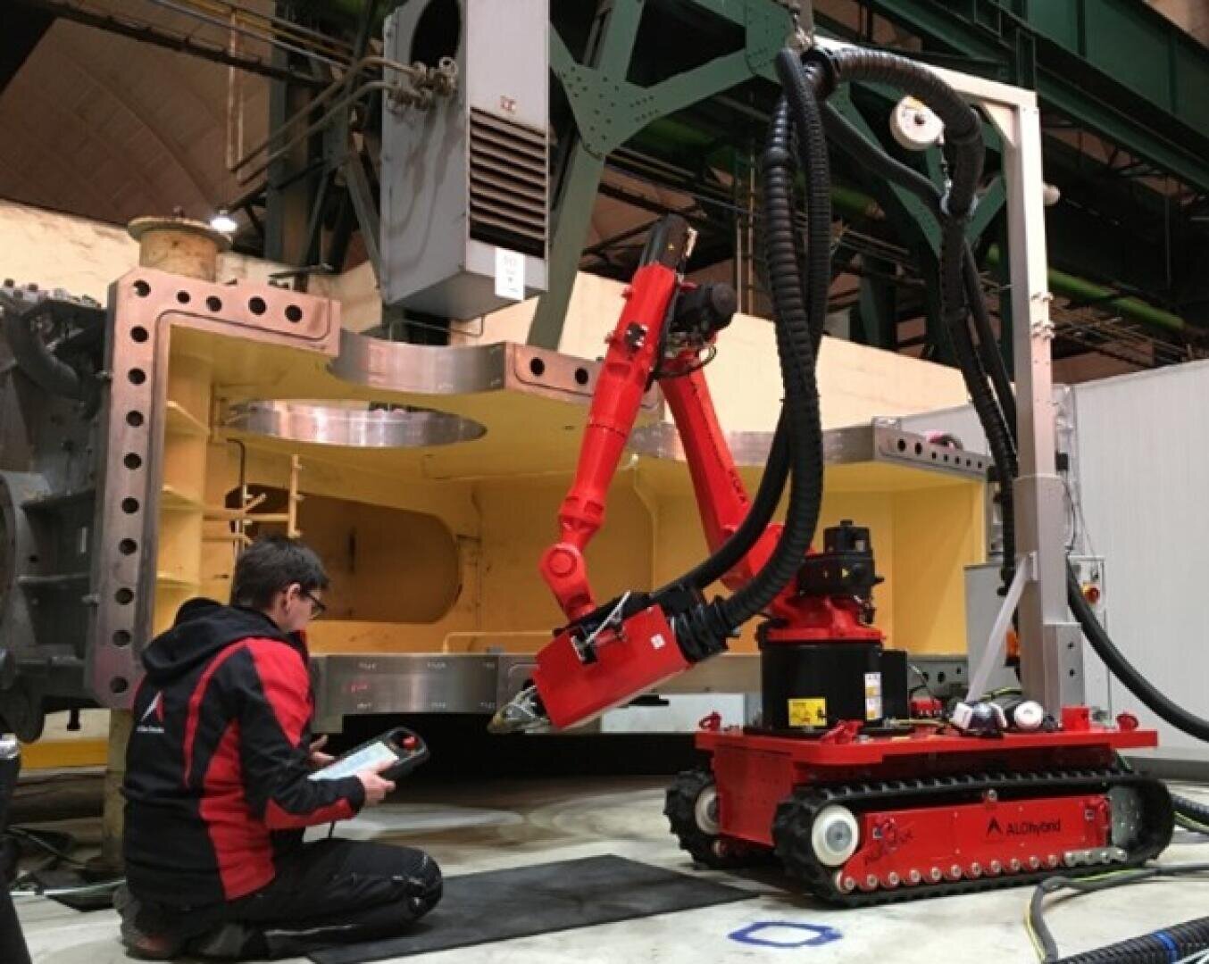 A person kneels on the ground, operating a remote control next to a red industrial robot with an extended arm, working inside a large metallic structure in an industrial facility.