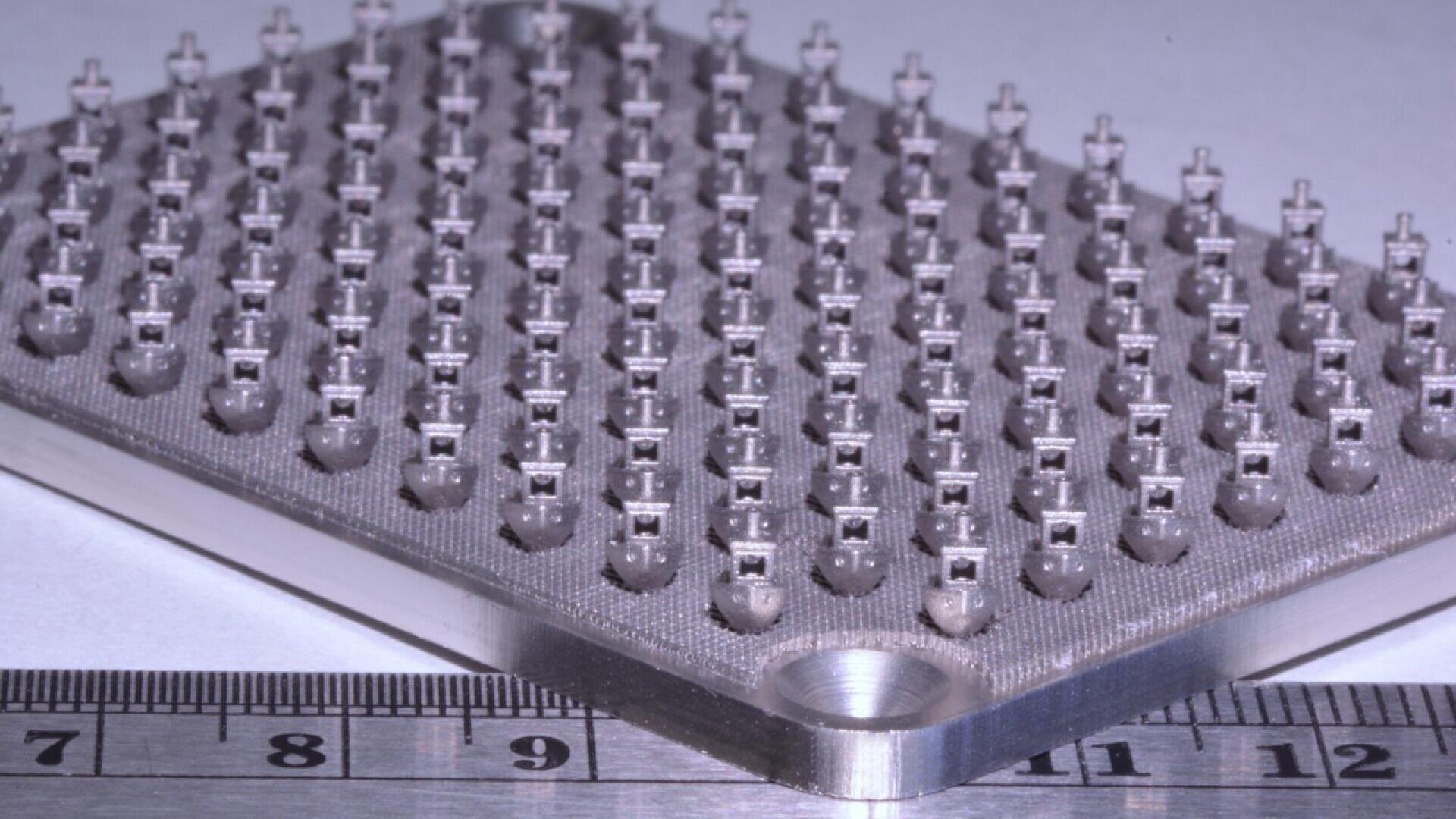 A metal plate with rows of small, precise cylindrical structures, shown next to a ruler for scale. The structures appear uniform and closely spaced, suggesting a high level of detail and precision.