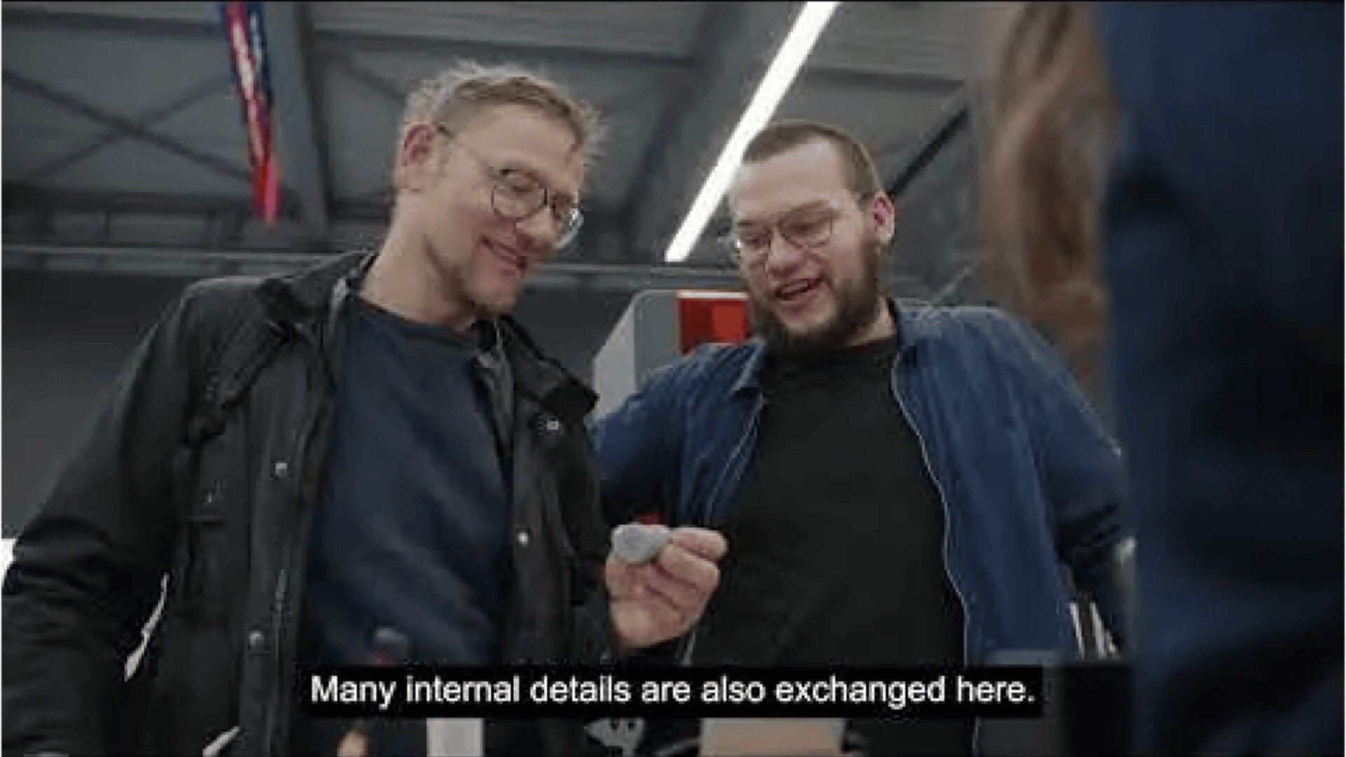 Two people stand close together, smiling and looking at an object one of them is holding. The setting appears industrial. Subtitle reads: “Many internal details are also exchanged here.”.