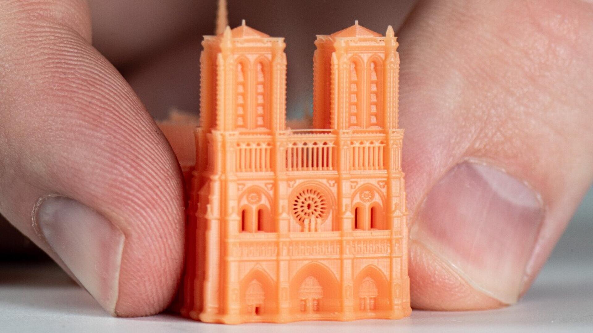 A close-up of fingers holding a small, orange 3D-printed model of the Notre-Dame Cathedral, showing detailed architectural features and twin towers.